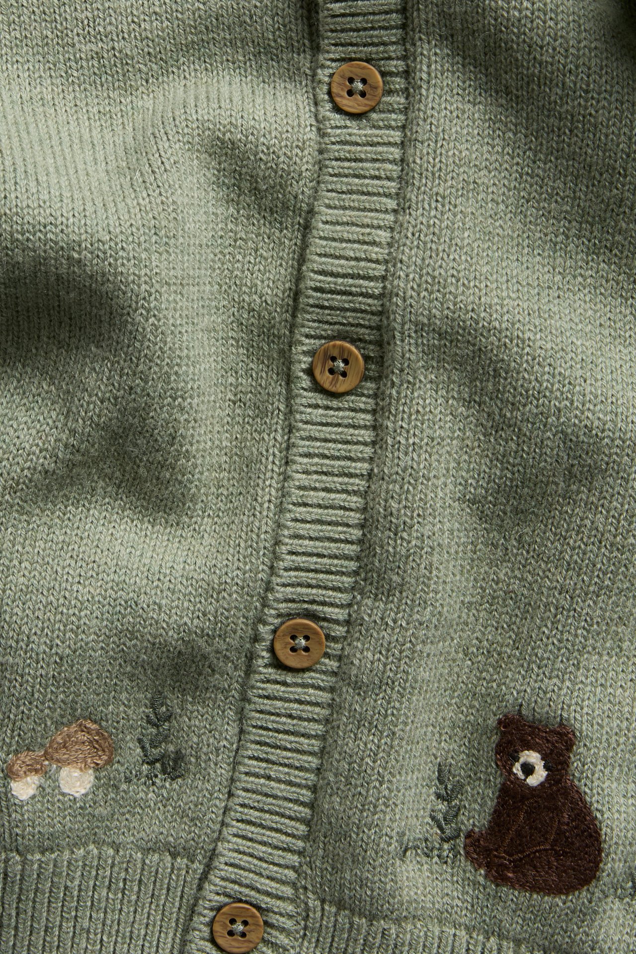 Nærbilde av en grønn strikket barnecardigan med brune knapper og broderte skogsdyr.