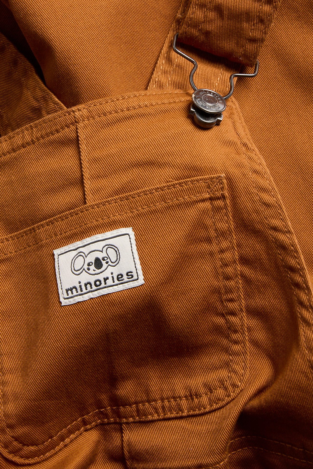 Närbild på mörk orange twillhängslebyxa med metallspänne och vit etikett med texten 'Minories'.