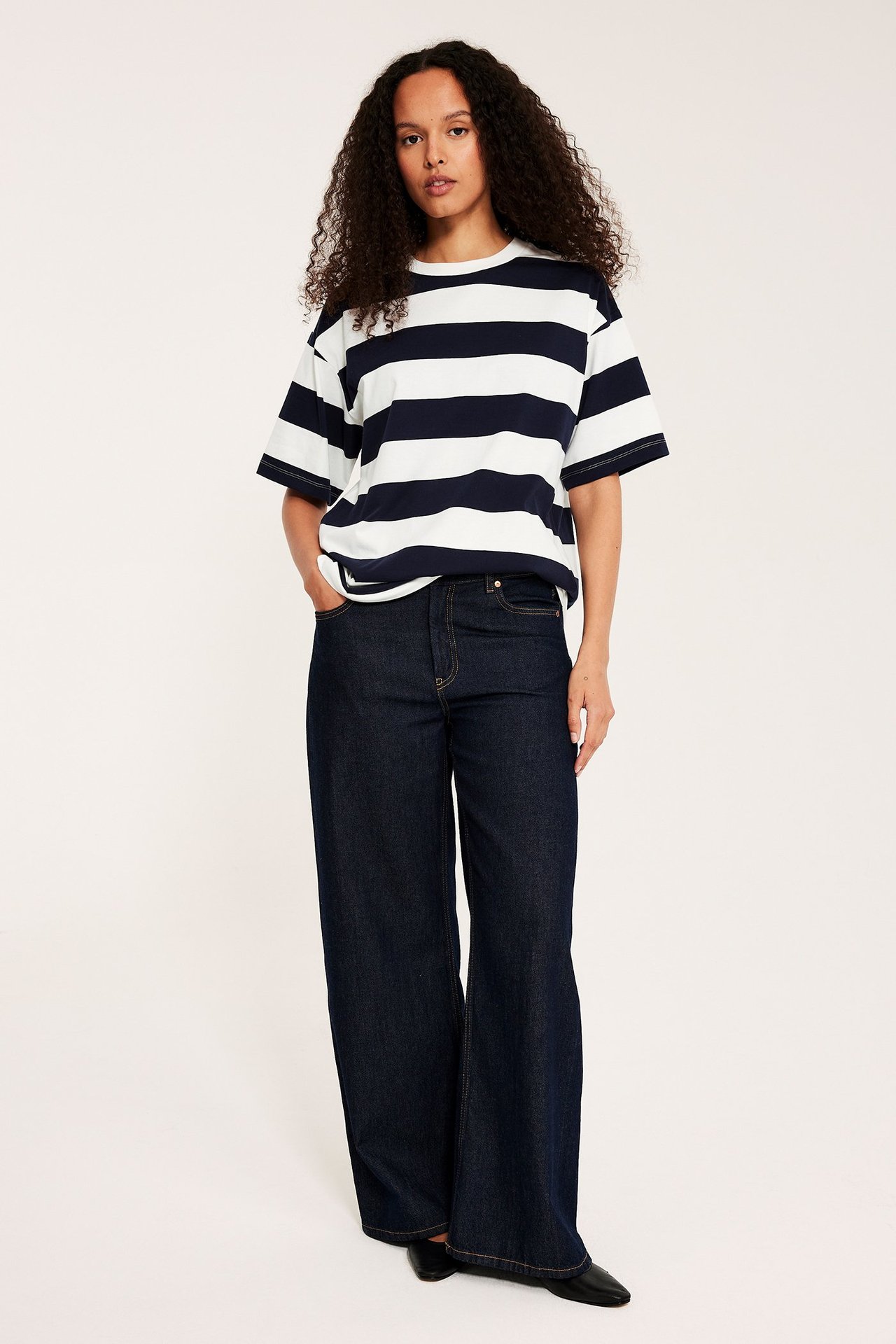 Forfra: kvinne i oversized mørkeblå og hvitstripet t-skjorte, mørkeblå vide jeans og svarte flate sko.