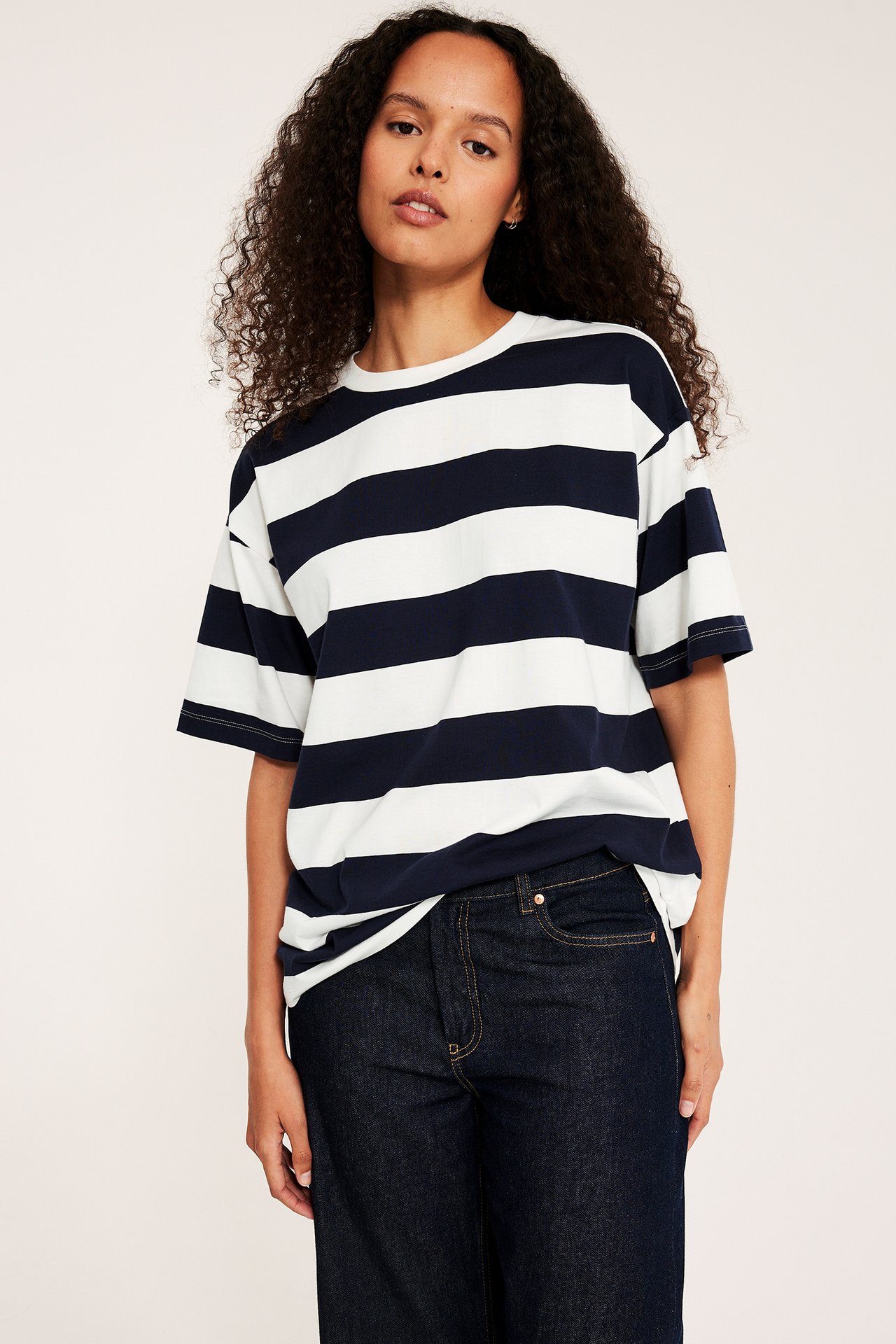 Naisen etunäkymä yllään ylisuuri laivastonsinivalkoraidallinen puuvillainen t-paita tummansinisten farkkujen kanssa.