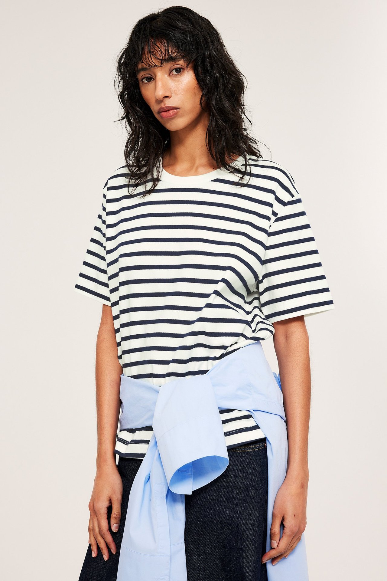 Nainen yllään oversized luonnonvalkoinen ja tummansiniraidallinen puuvilla-t-paita. Vaaleansininen paita on sidottu vyötärölle.