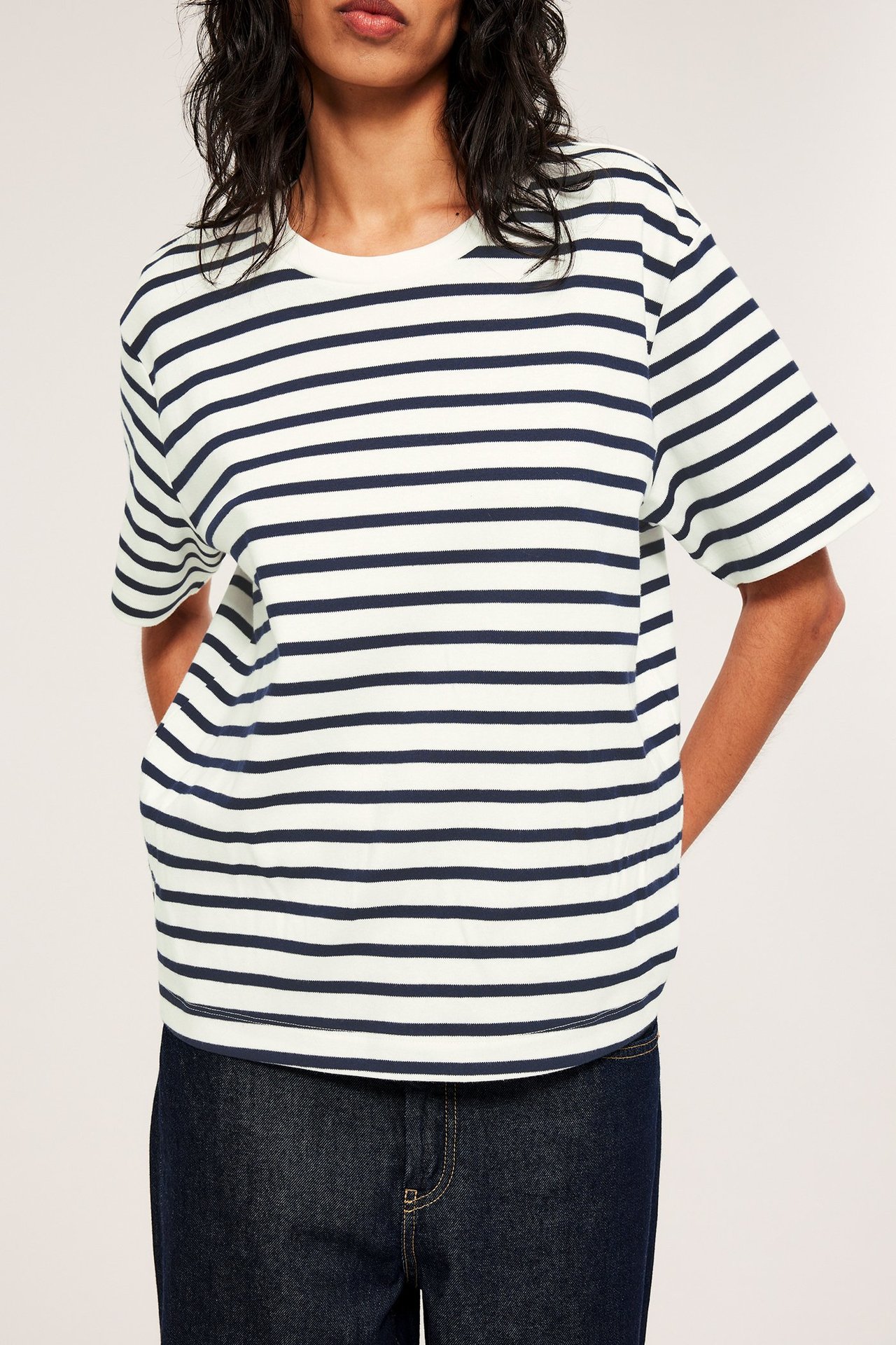 Edestäpäin kuvassa nainen yllään oversized luonnonvalkoinen puuvillainen t-paita tummansinisillä raidoilla ja tummansiniset farkut.