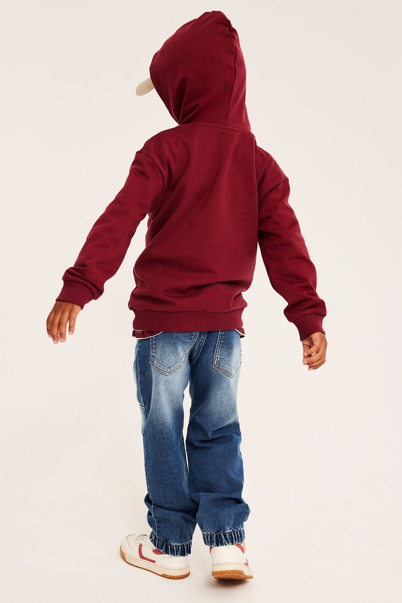 Ryggvy av barn i vinröd huvtröja, blå jeans, vita sneakers och beige keps.
