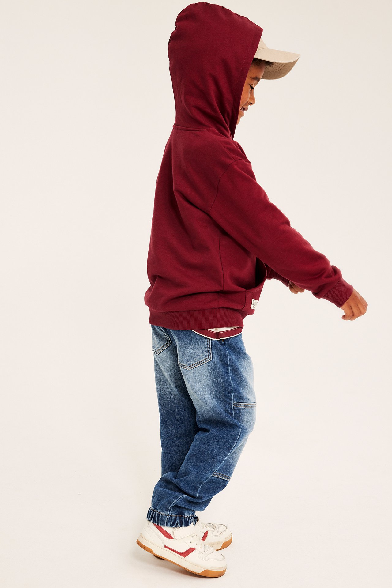 Barnets sidovy i vinröd hoodie, blåa denimbyxor, vita sneakers med rött och en beige keps.