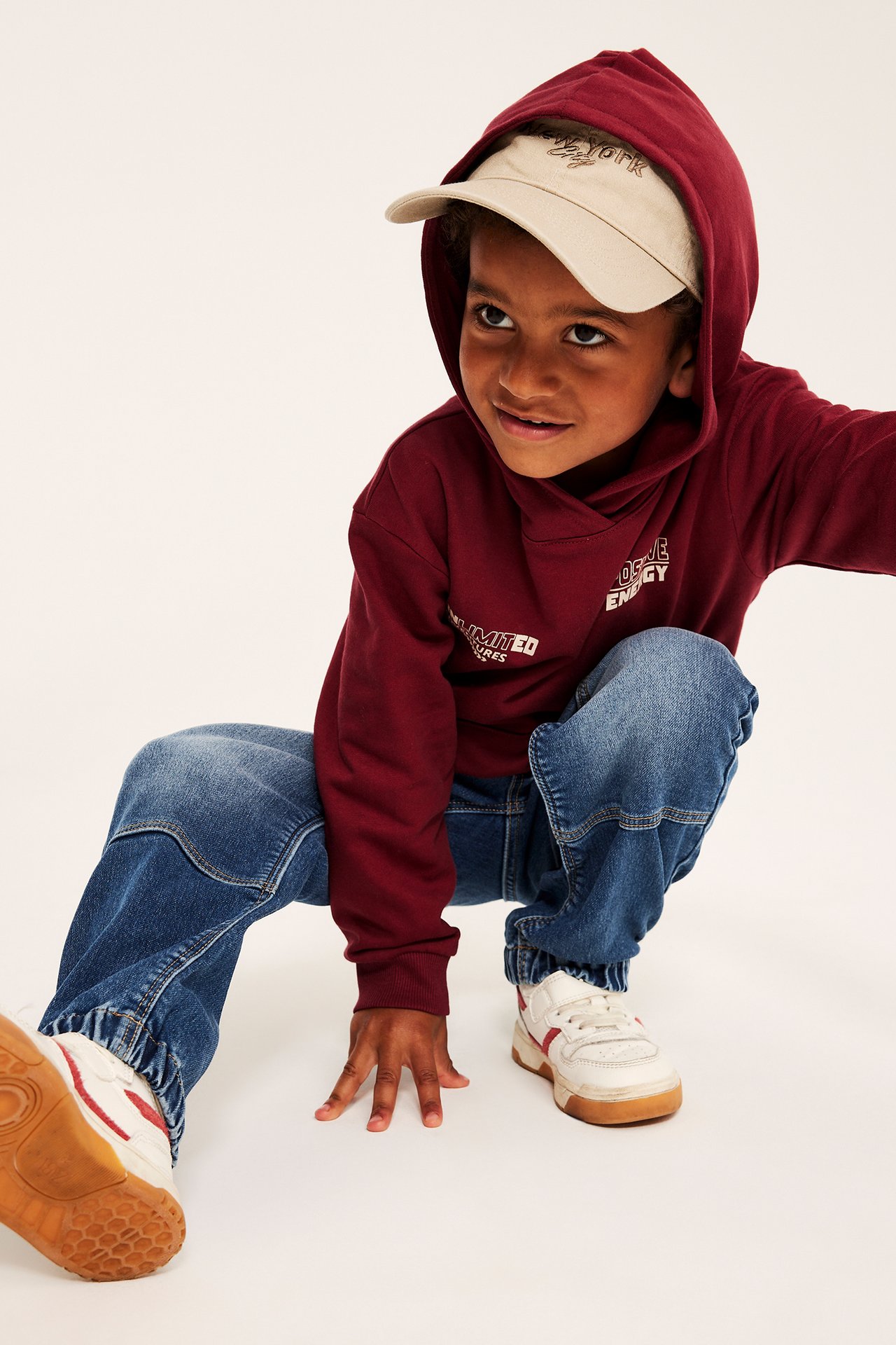 Framifrån, ett barn i en vinröd hoodie med vit text, blå jeans, beige keps och vita sneakers.