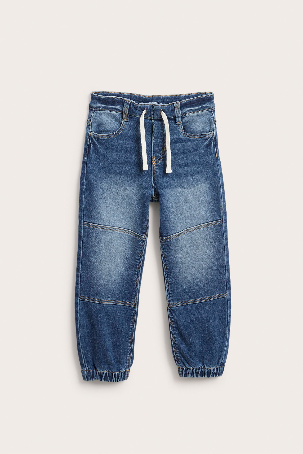 Framifrån: Blåa denimjoggers för barn med vita dragsko och knäpaneler.