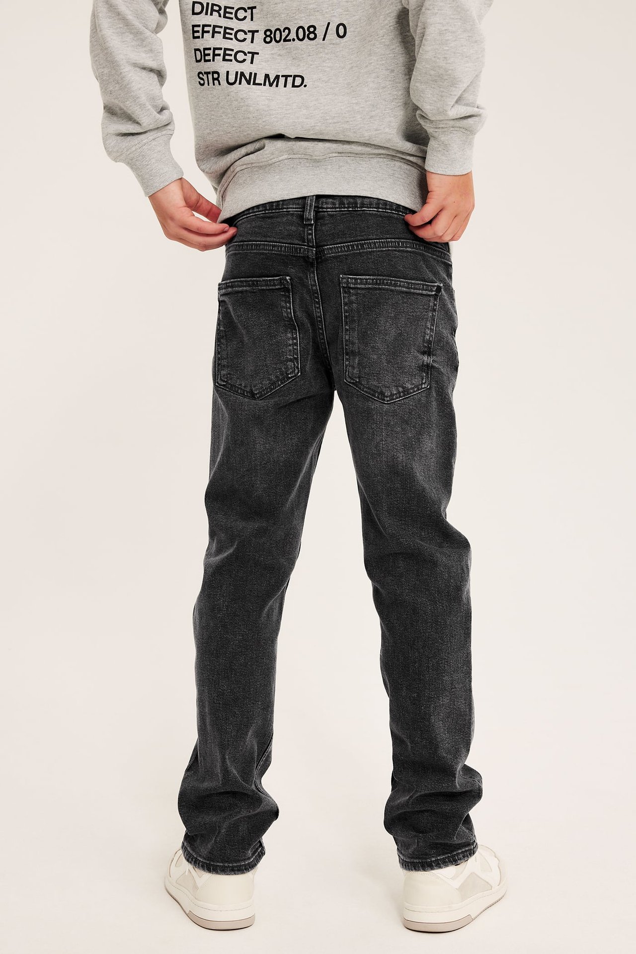 Bakfra av et barn iført mørkegrå vaskede jeans med rett passform, en grå hettegenser og hvite joggesko.