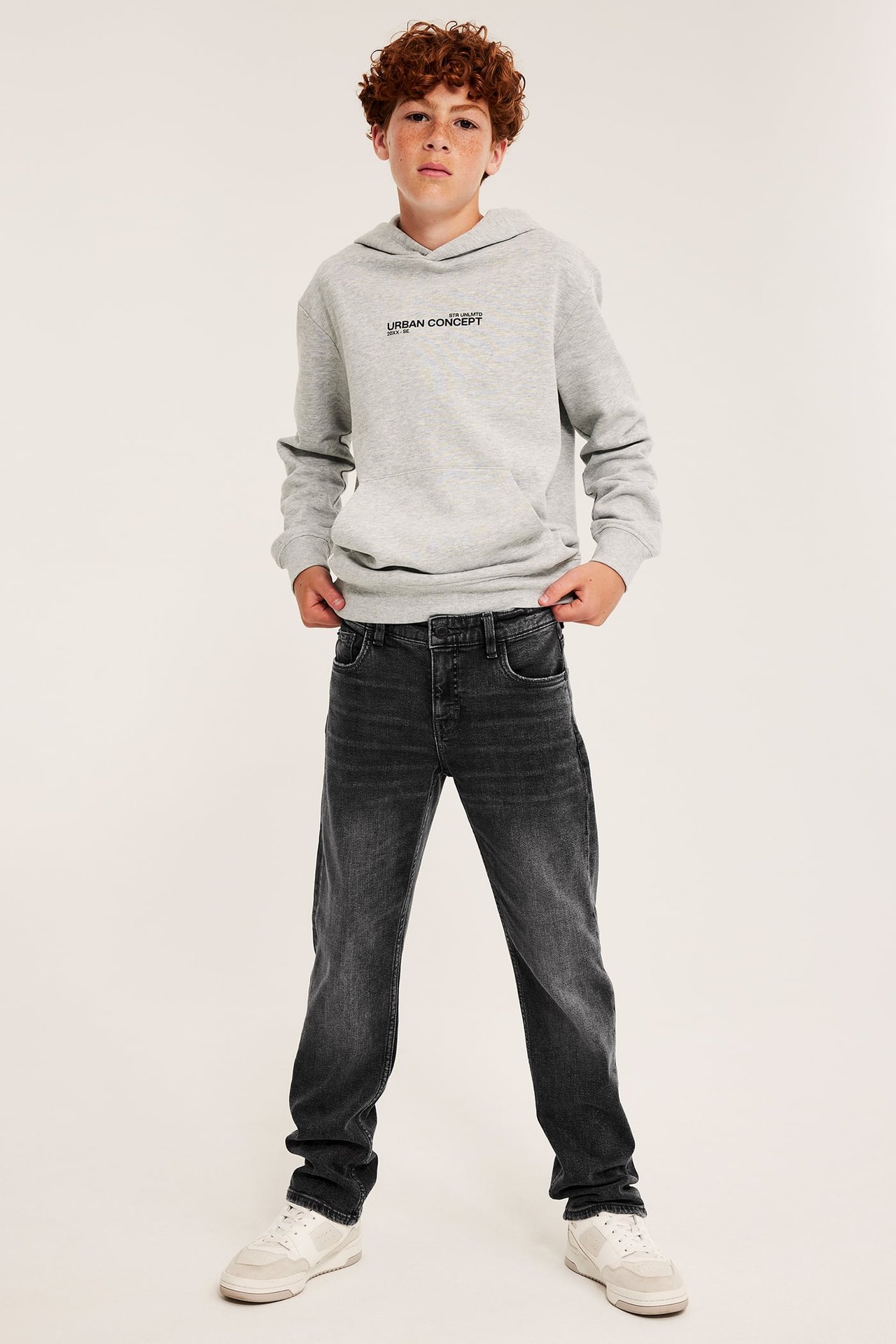 Front view av en gutt i grå hettegenser med svart trykk, svarte jeans og hvite joggesko.