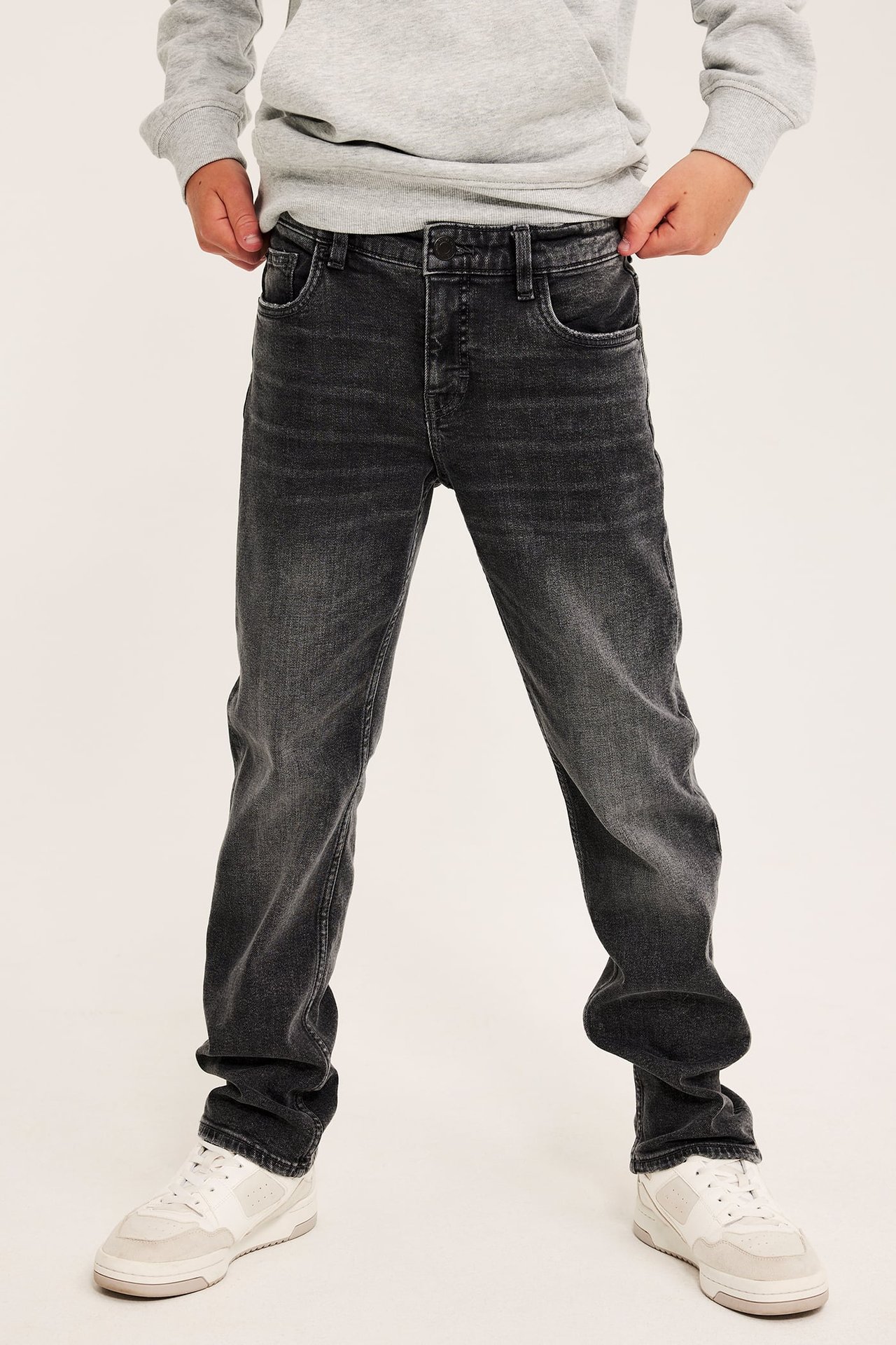 Forfra: et barn i vaskede sorte rette jeans, en grå genser og hvite joggesko.