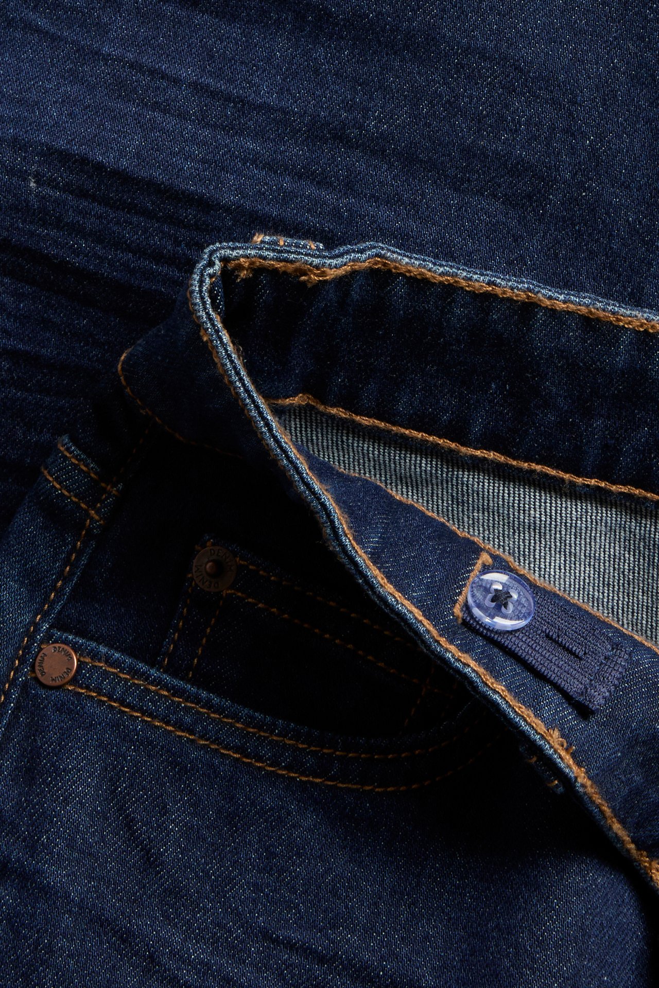 Närbild av mörkblå denimjeans med brun söm och justerbar midja med knapp.