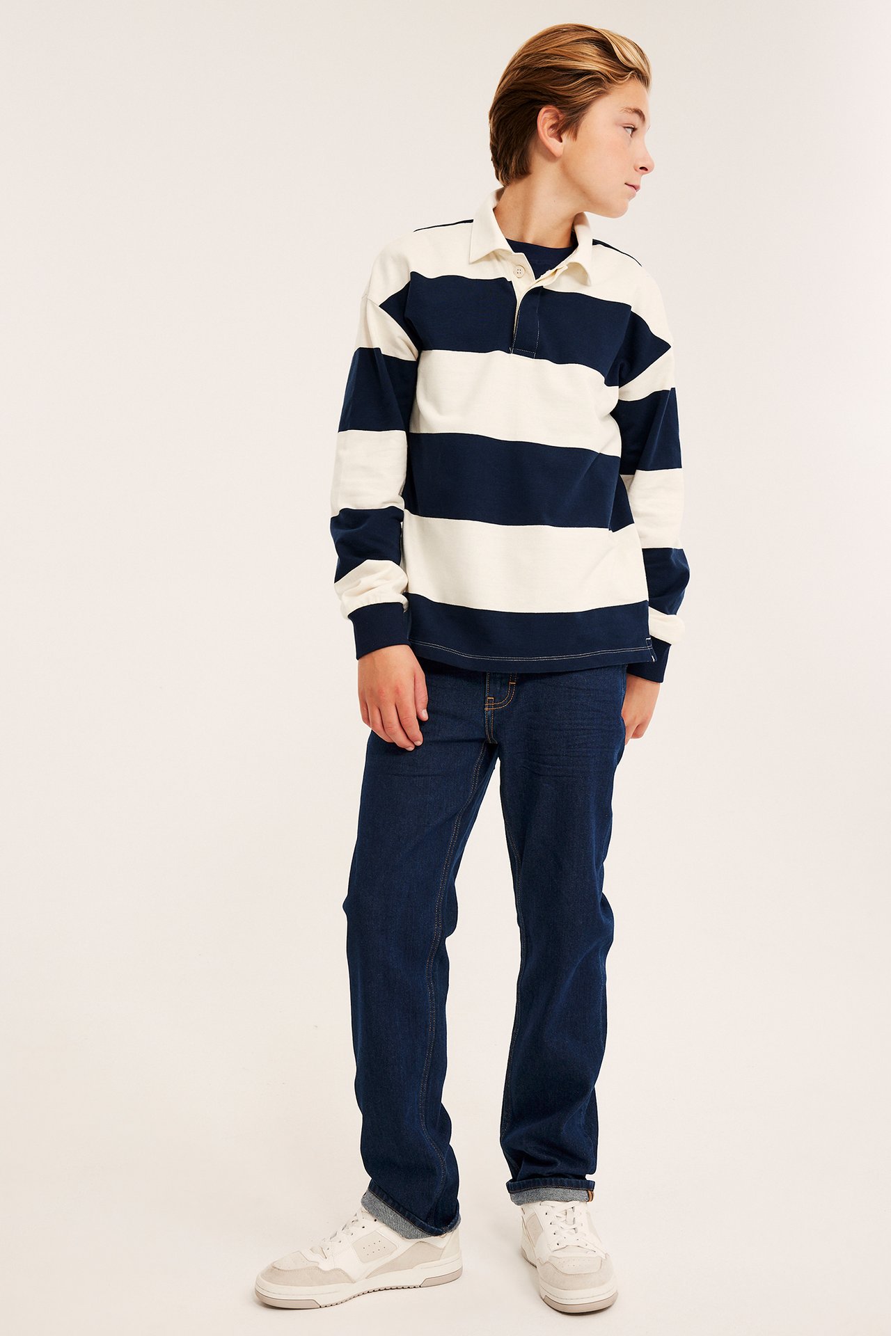 Front av barn med marineblå og offwhite stripet rugbyskjorte, mørkeblå jeans og hvite joggesko.