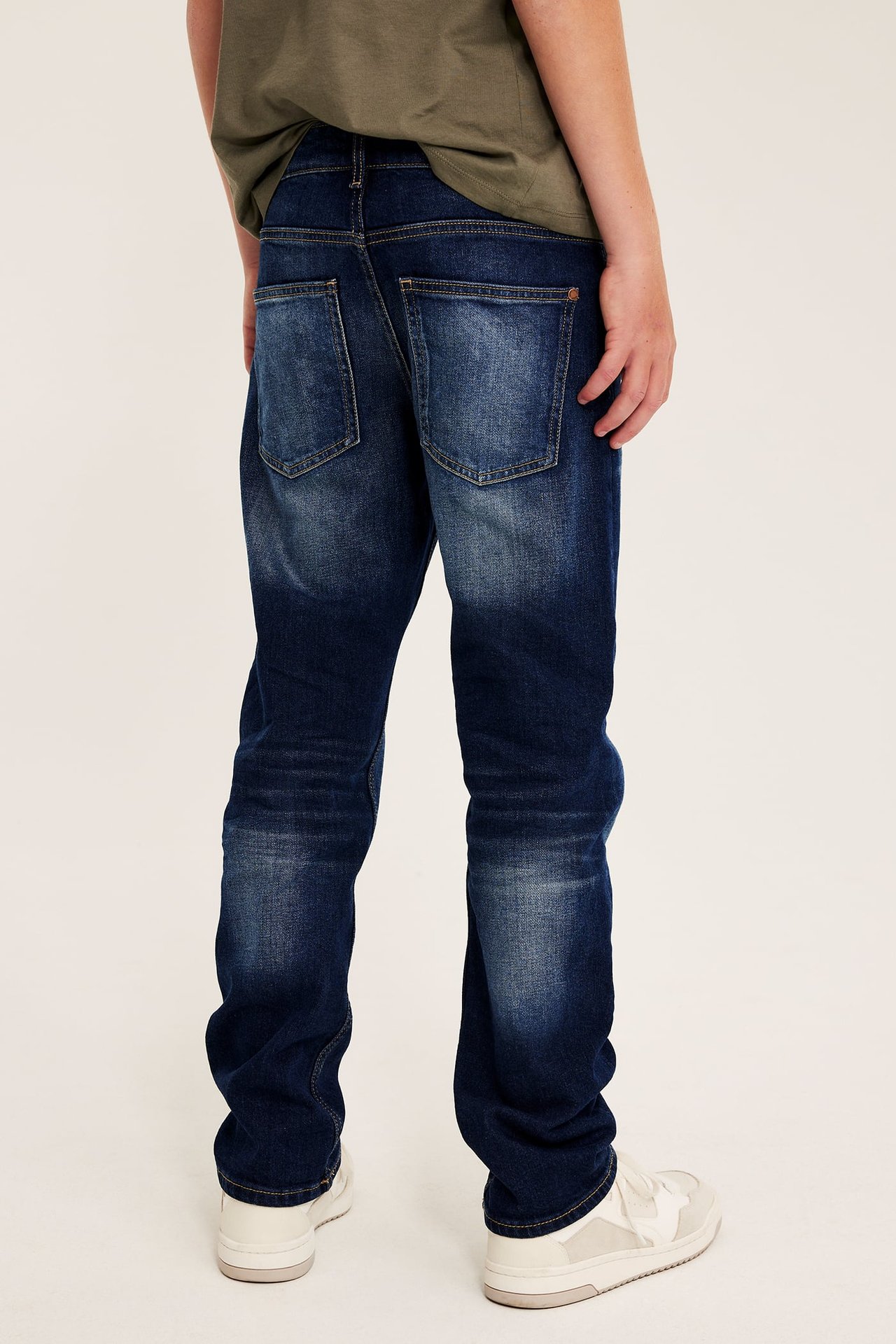 Bakfra bilde av et barn i mørkeblå rette jeans, grønn t-skjorte og hvite joggesko.