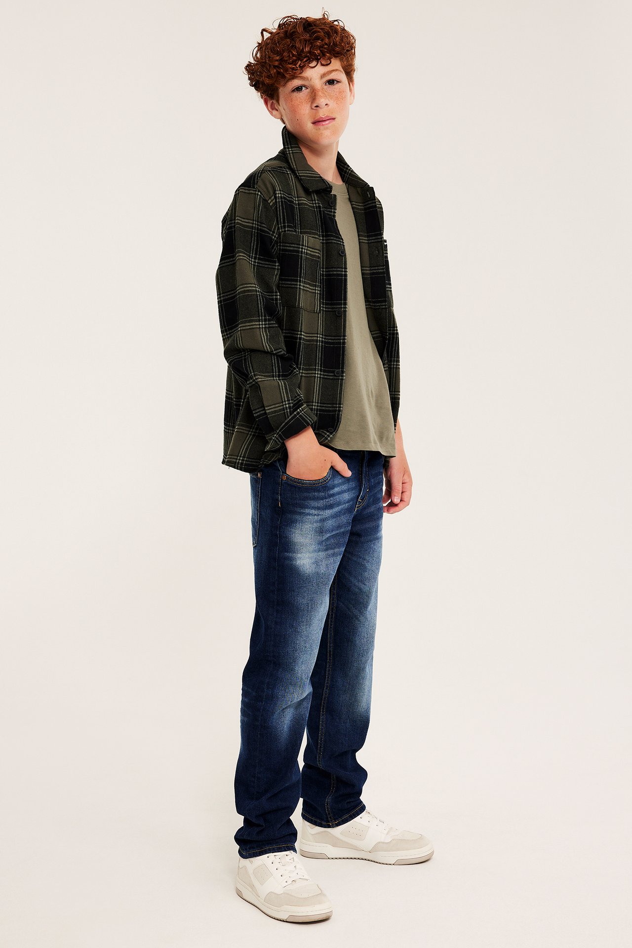 Sidovy av en pojke i en mörkgrön rutig skjorta, olivfärgad t-shirt, blå jeans och vita sneakers.