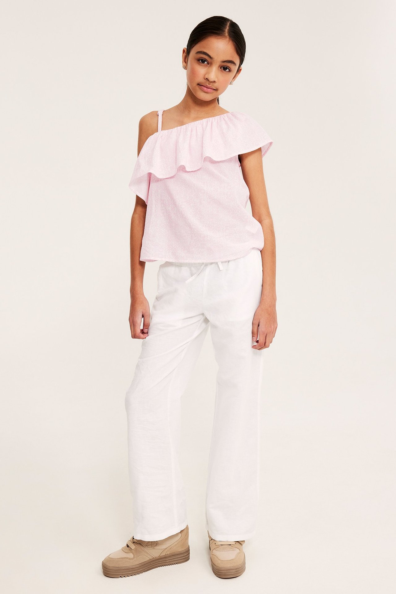 Frontbilde av et barn med en rosa mønstret one-shoulder volangtopp, hvite bukser og beige sneakers.