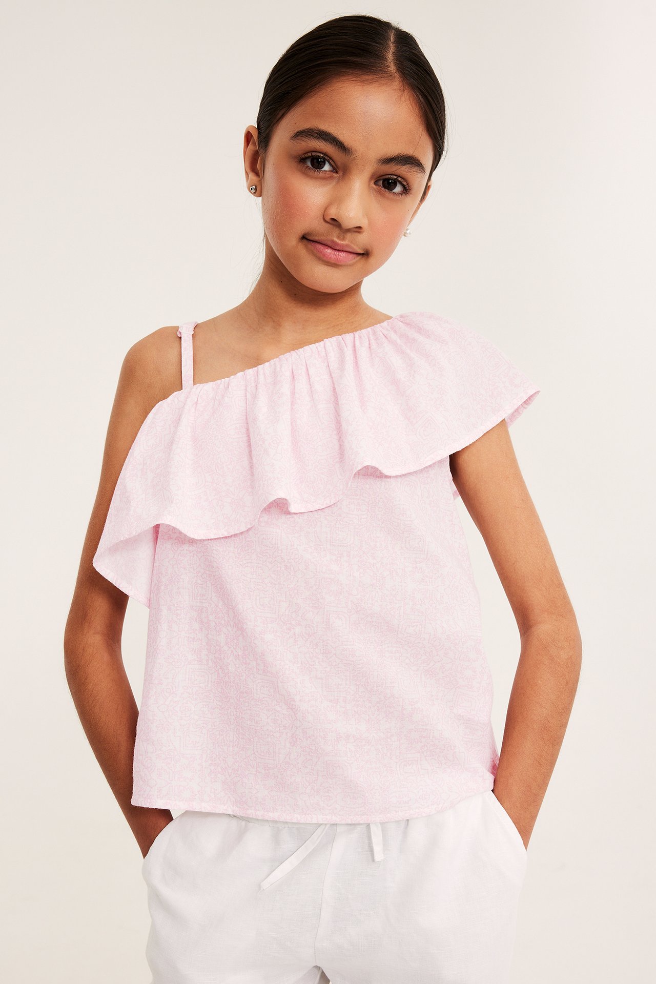 Framifrån, ett barn i rosa mönstrad one-shoulder topp med volang och vita shorts.