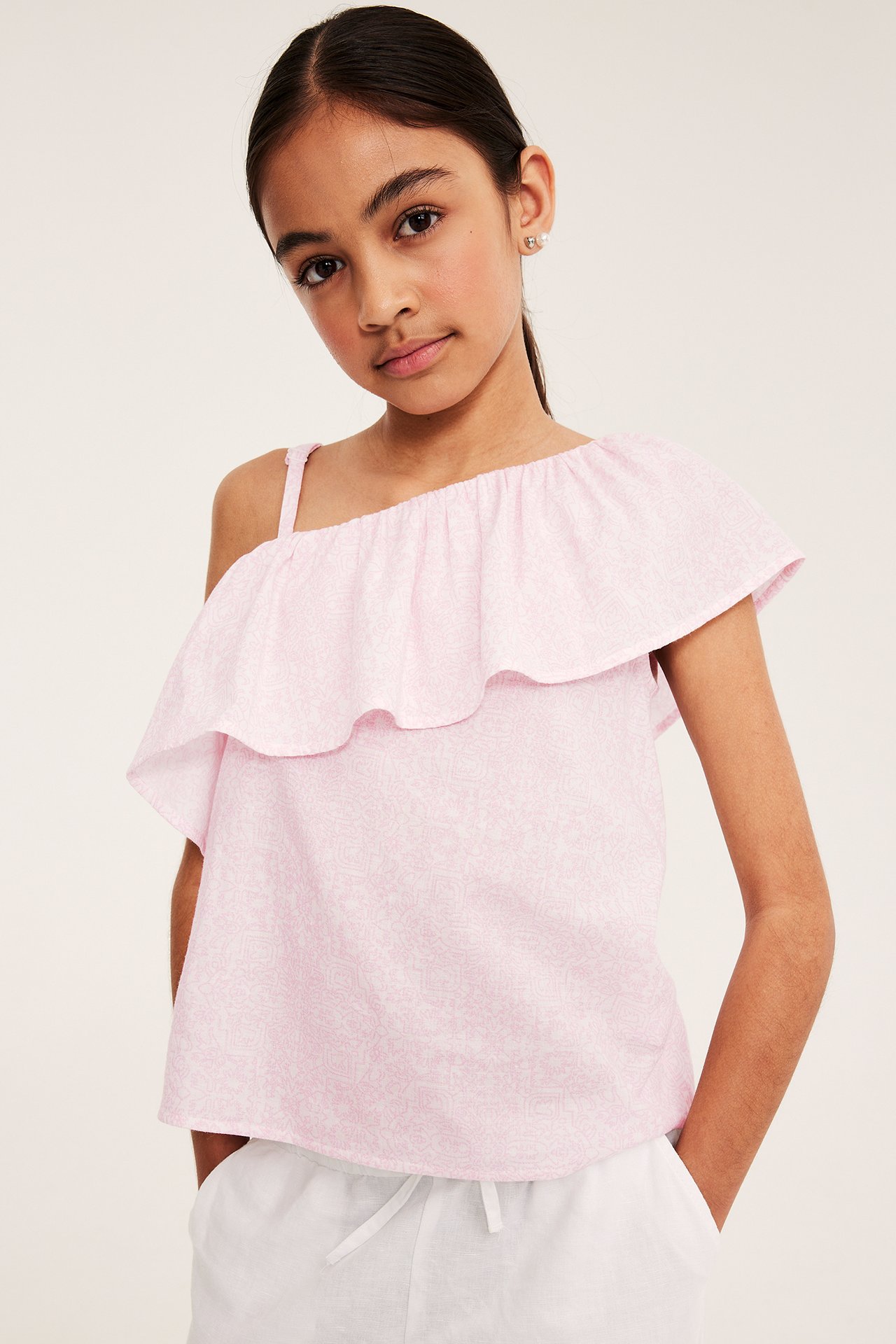 Framifrån, ett barn i en ljusrosa mönstrad one-shoulder bomullstopp med volanger, matchad med vita byxor.