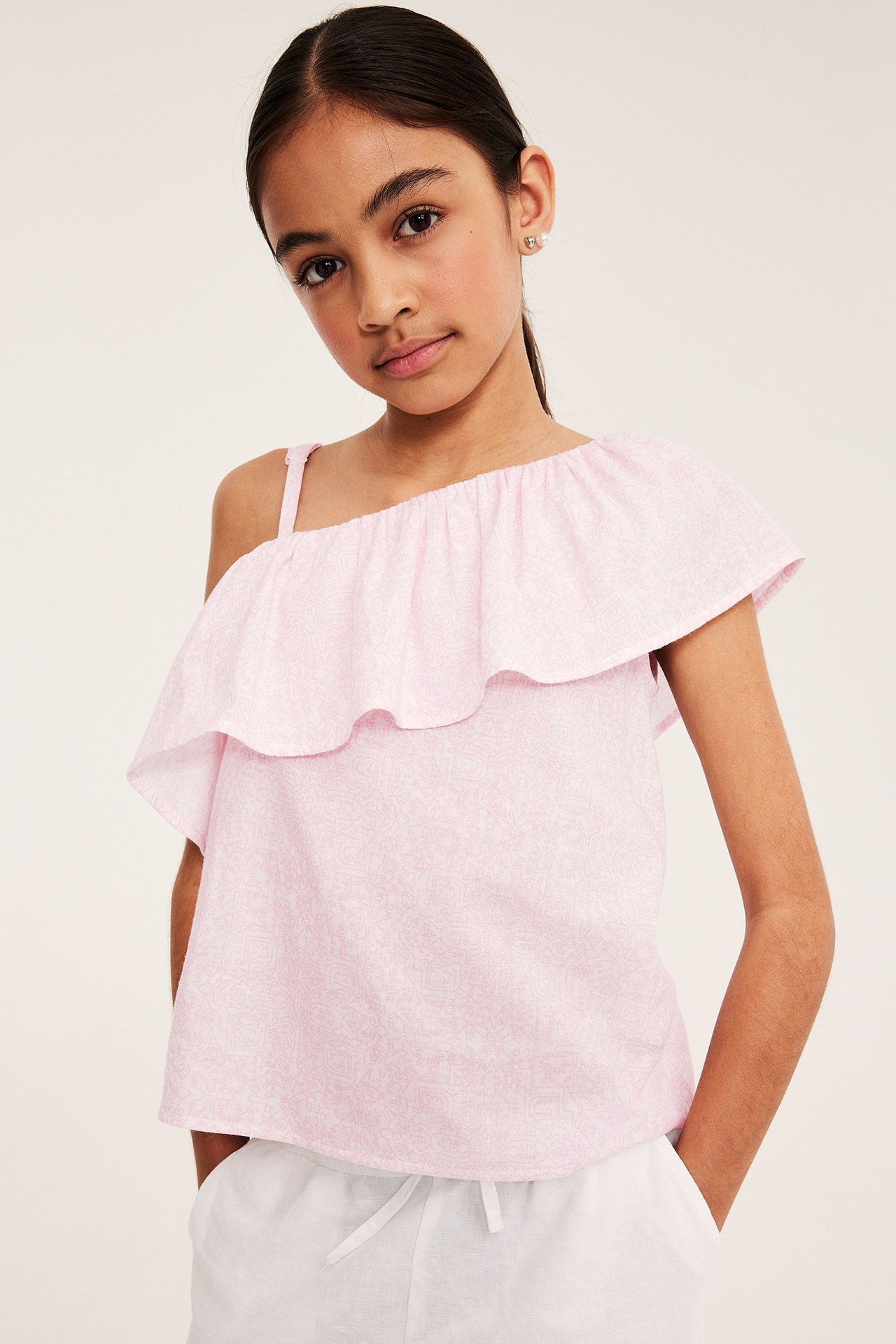 Widok z przodu na dziecko w różowym wzorzystym bawełnianym topie na jedno ramię z falbankami i białymi spodniami.