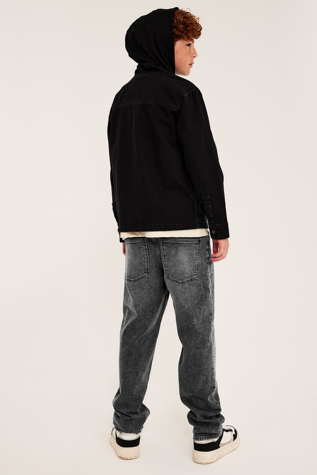 Bakfra av et barn i svart hettejakke i denim, grå acid wash-jeans og svarte og hvite joggesko.