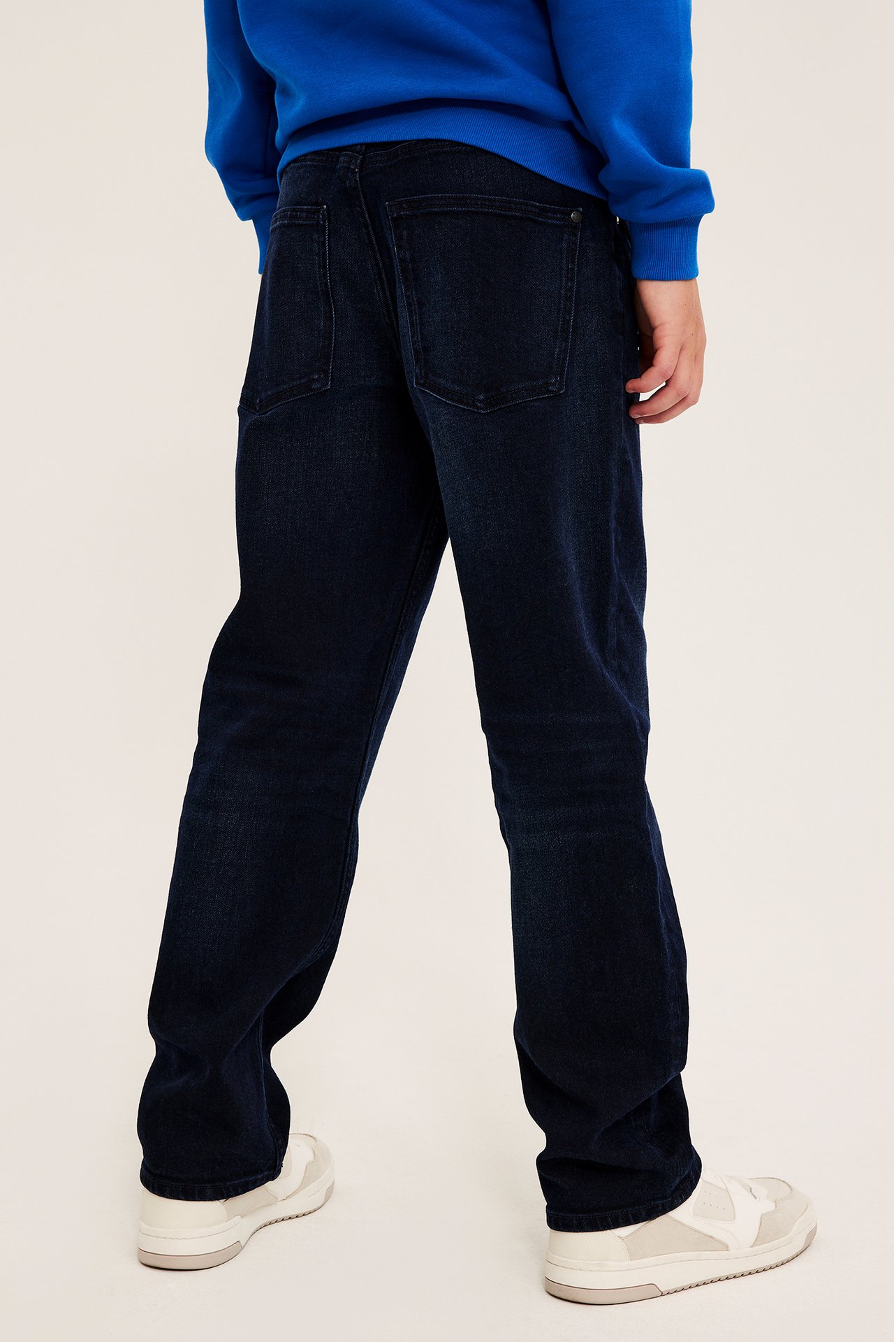 Bakfra av person i mørkeblå rette jeans, kongeblå genser og hvite joggesko.