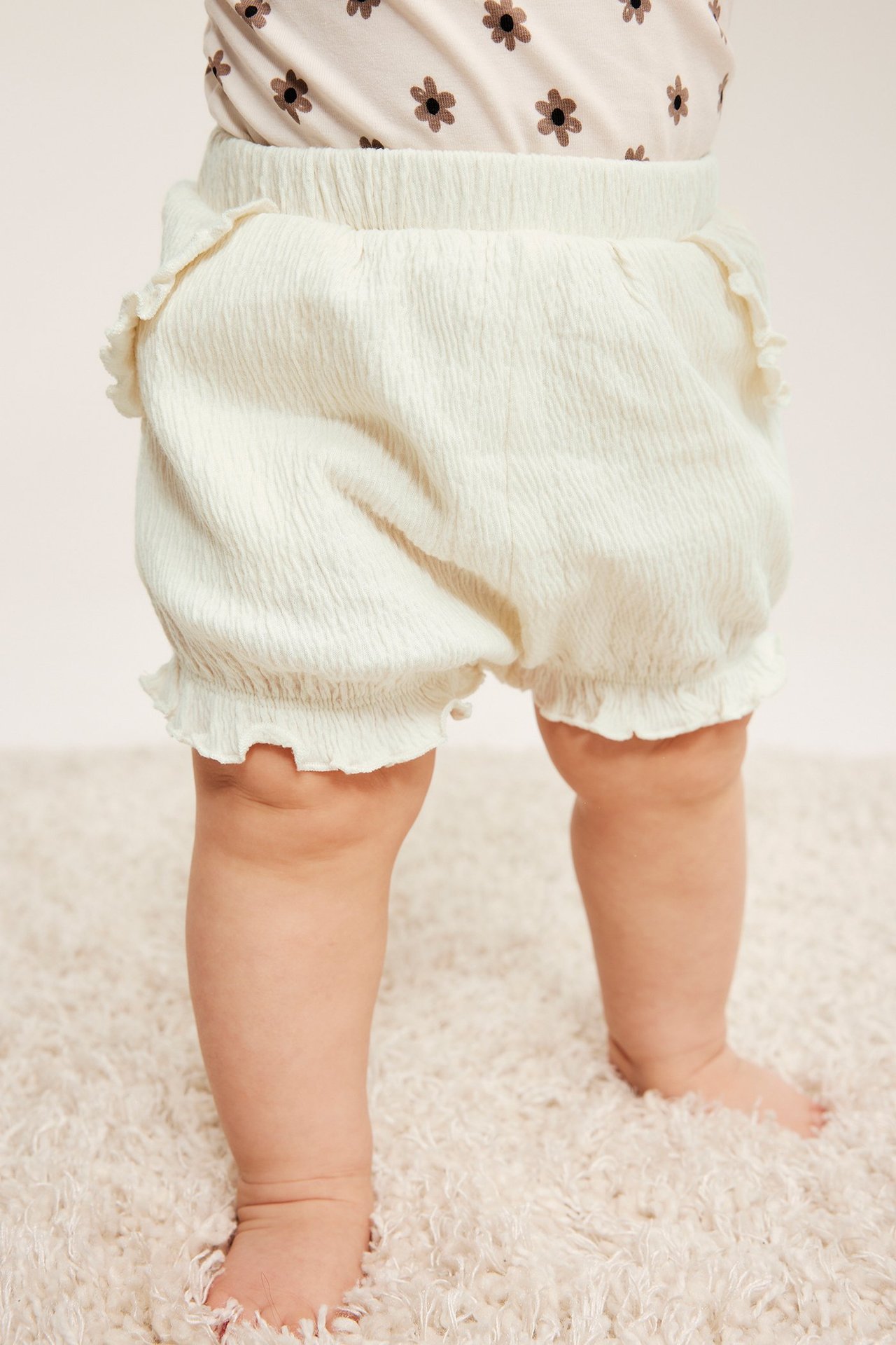 Baby i kremhvite rynkede bomullsshorts med volanglommer, elastiske ben og blomstertopp.