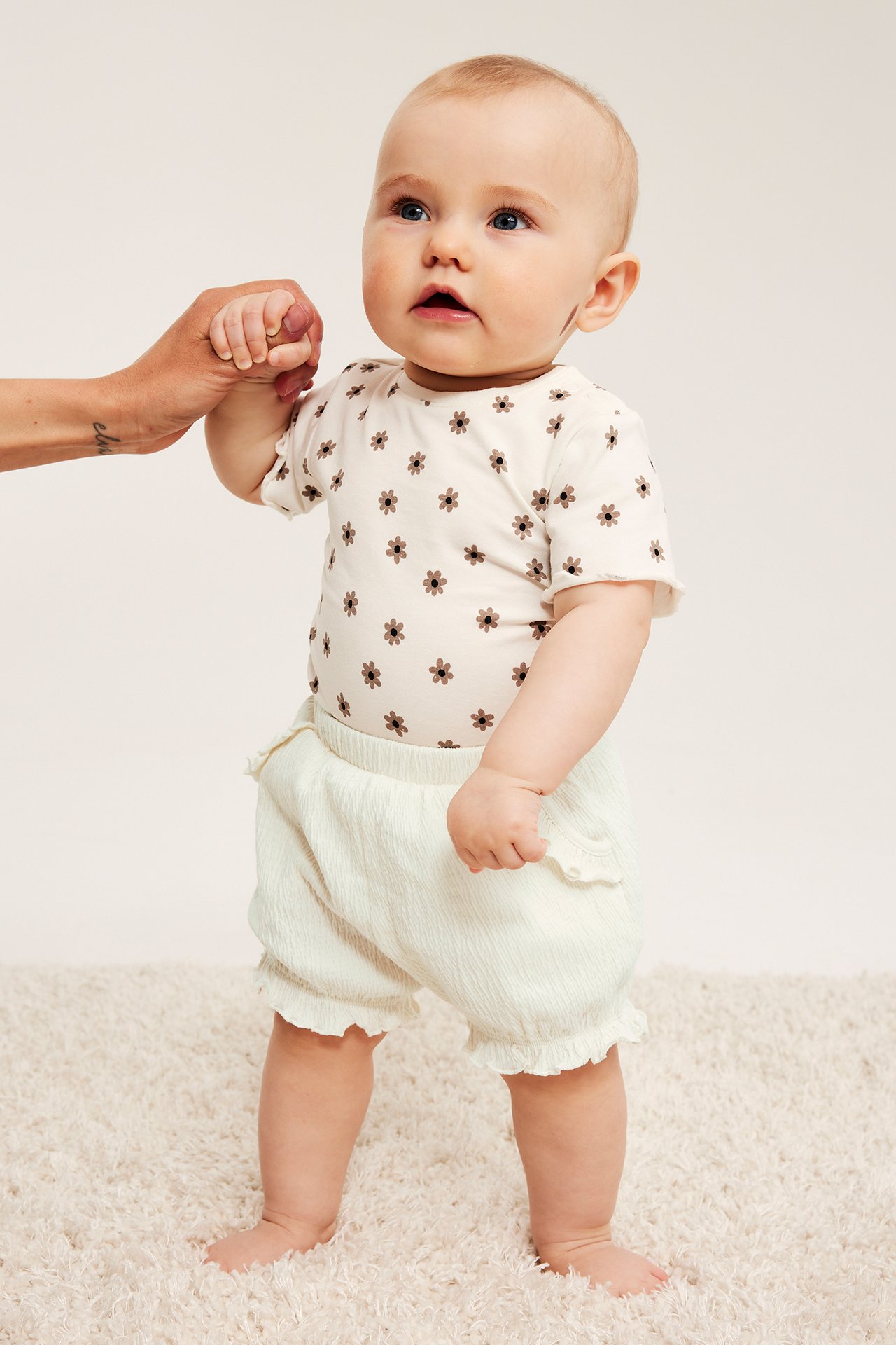 Framifrån: baby i benvita krinklade shorts med volanger, blommig body, håller en vuxens hand.