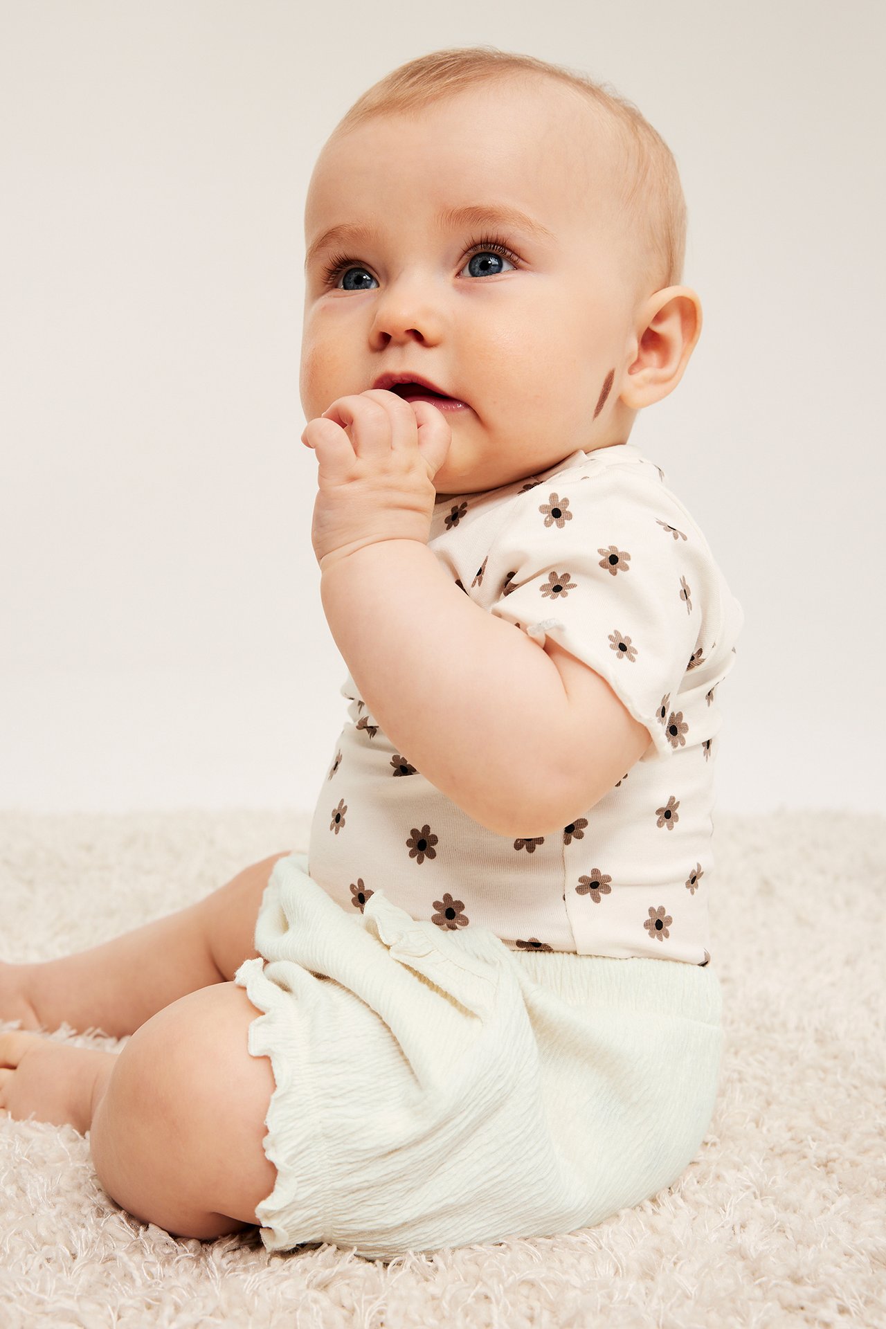 Widok z boku niemowlaka w off-white marszczonych bawełnianych szortach z falbankami i kwiecistym off-white body.