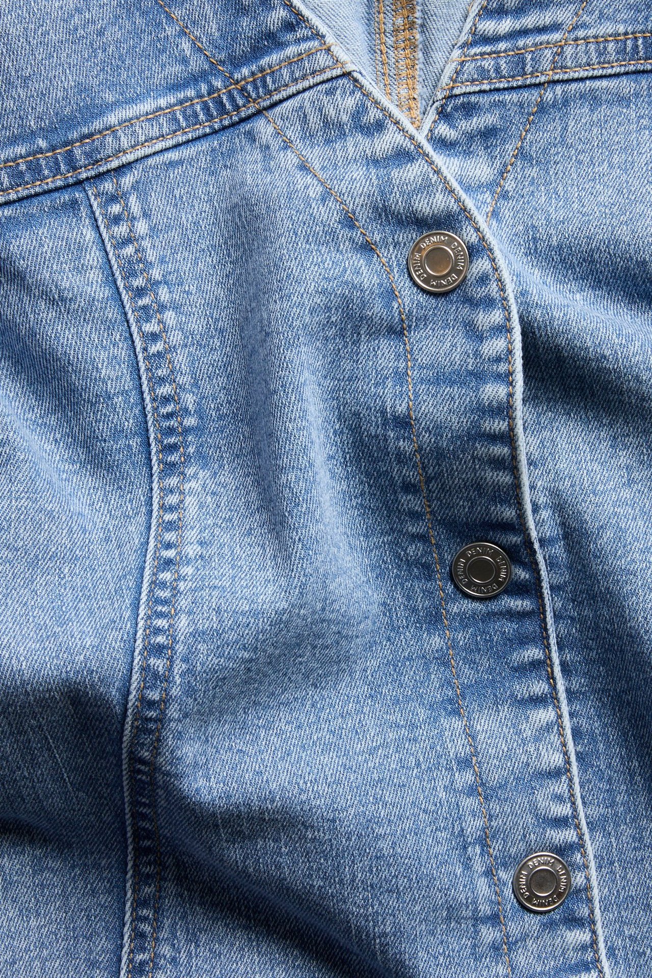 Zbliżenie jasnoniebieskiej dopasowanej kurtki-bluzy jeansowej, z jasnobrązowymi szwami i srebrnymi guzikami.