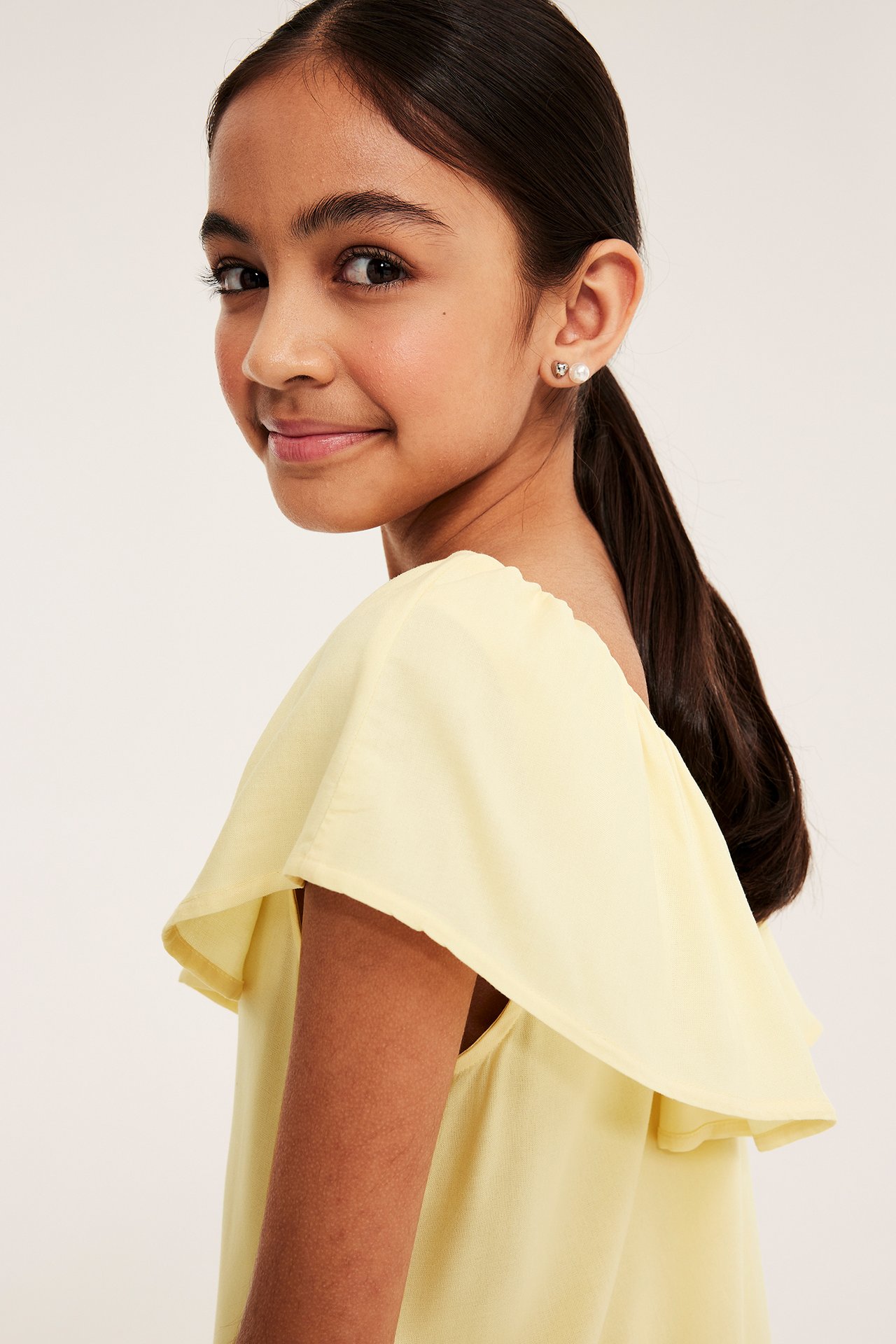 Sivukuva lapsesta vaaleankeltaisessa, röyhelöreunaisessa viskoosimekossa ja helmikorvakoruissa.