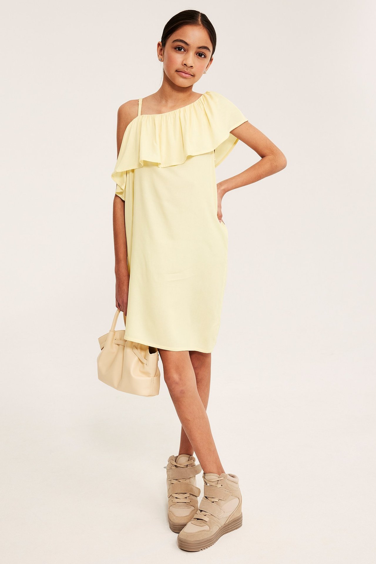 Edestä, lapsi pukeutunut vaaleankeltaiseen yksihihaiseen viskoosimekkoon röyhelöillä, pitää beigen laukkua ja tennareita.