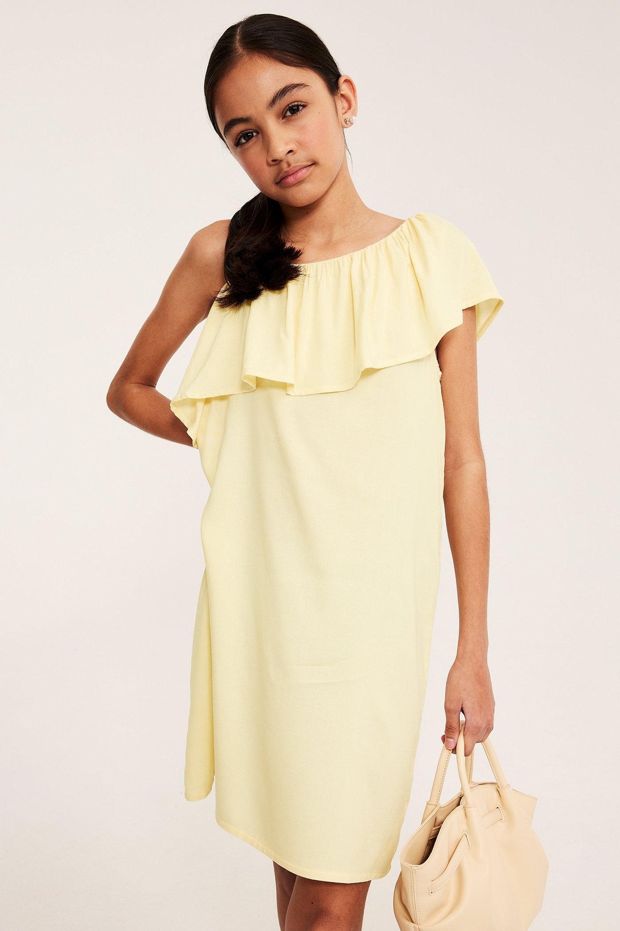 Lapsi edestä: vaaleankeltainen viskoosimekko yhdellä olalla ja röyhelöllä, pitää beigen käsilaukkua.