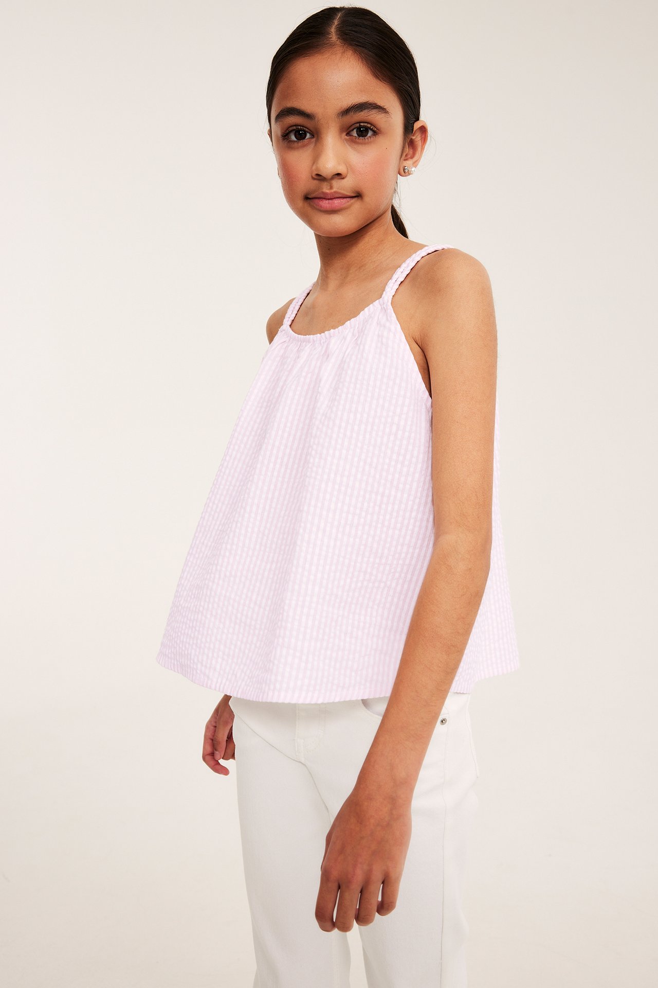 Barn bär en lös rosa randig bäckebölja ärmlös topp med flätade axelband och vita byxor.