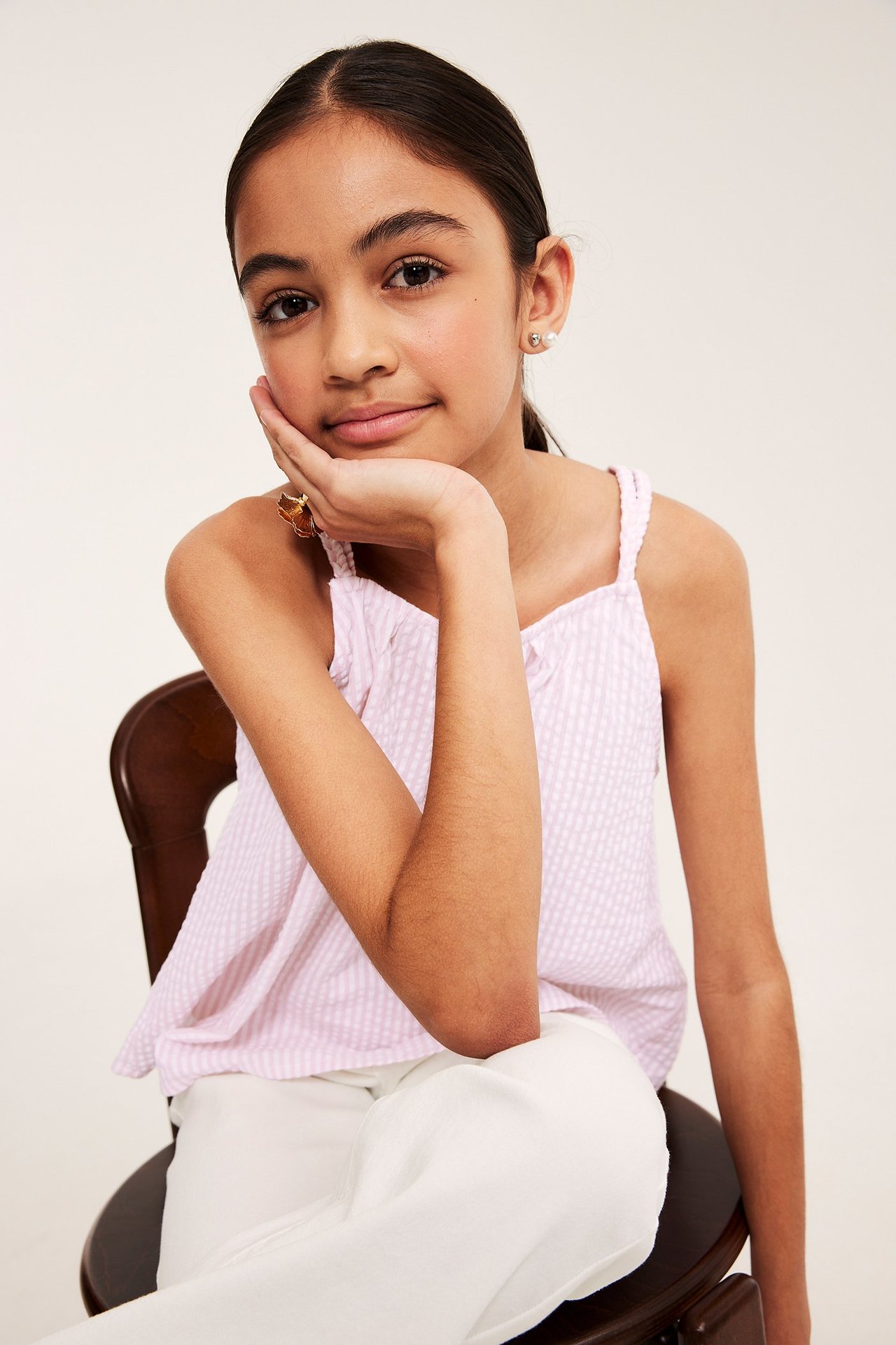 Frontvy av ett barn i rosa randig ärmlös topp med flätade band, ljusa byxor, ring och örhängen.