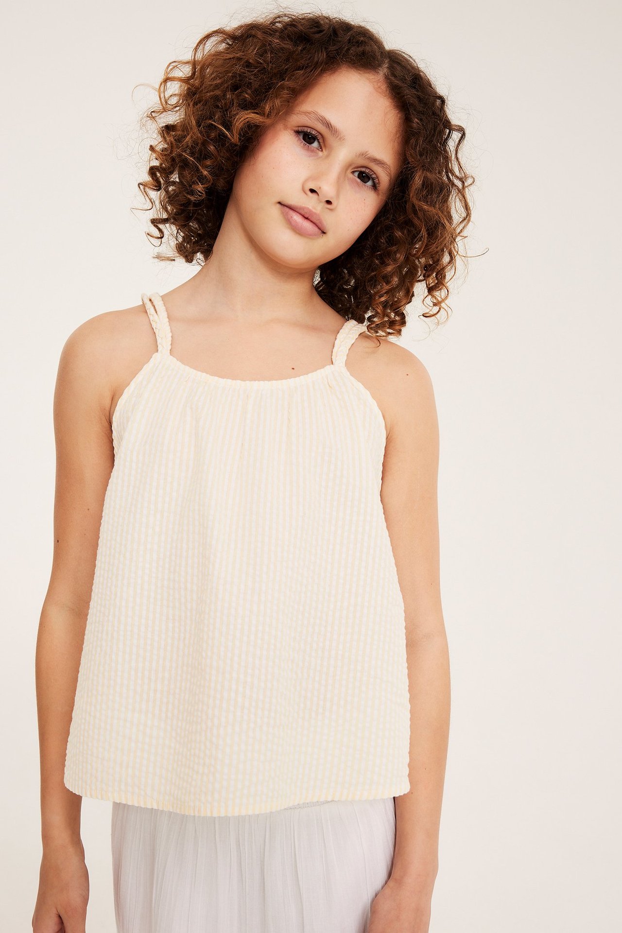 Frontvy: Ett barn bär en gulrandig ärmlös topp i bäckebölja med flätade axelband och grå kjol.