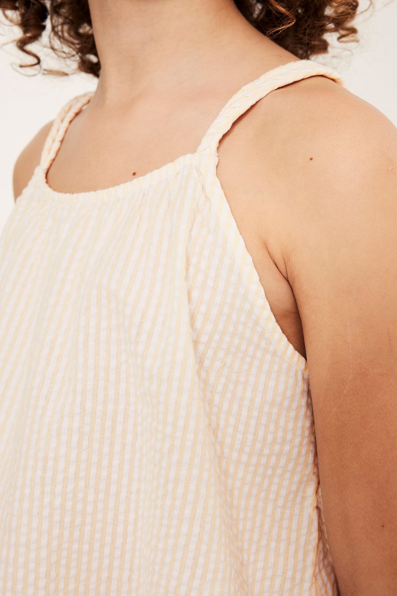 Närbild, sidovy: barn i gul-vitrandig ärmlös bomullstopp i bäckebölja med flätade axelband.