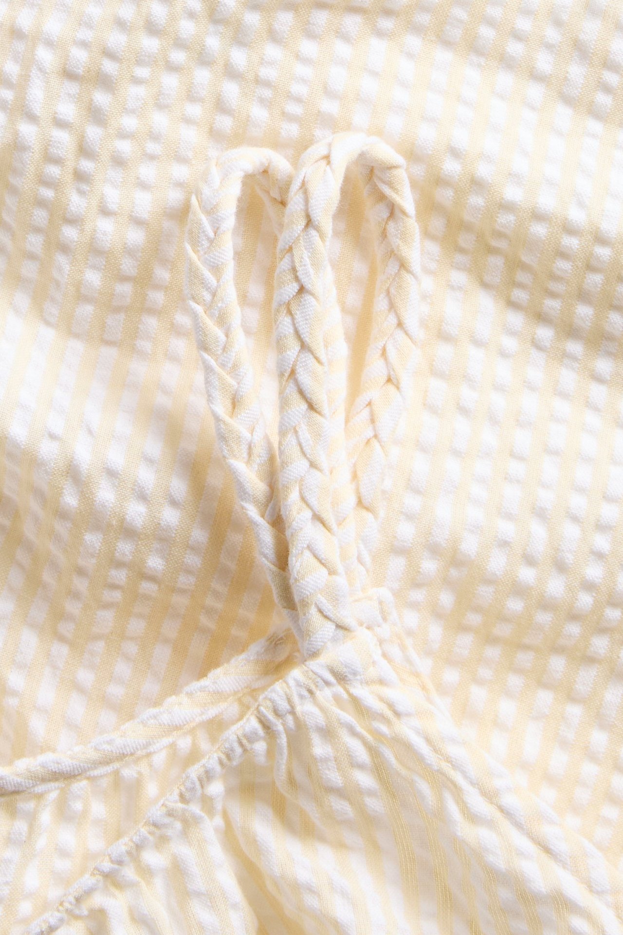 Närbild av en gulvit randig ärmlös bomullstopp i bäckebölja med flätade axelband och rynkad kant.