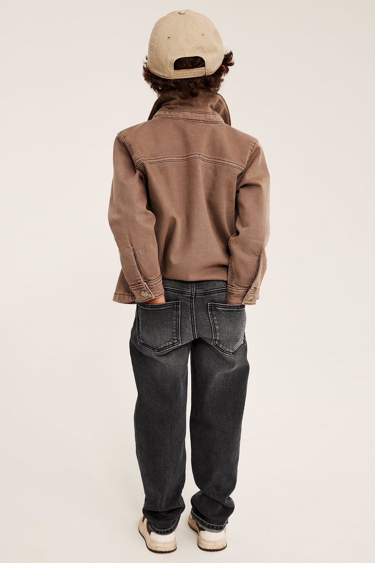 Bakifrån: Barn i brun överskjorta, svarta jeans, beige keps och sneakers.