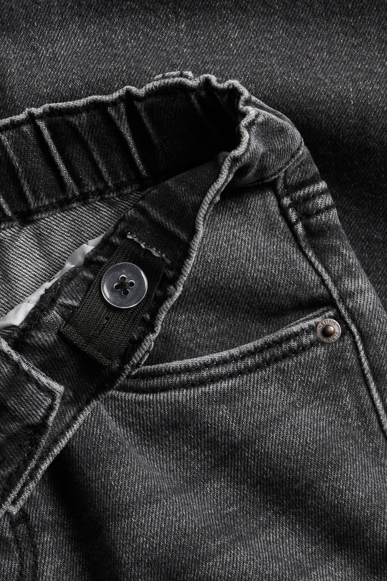 Närbild på mörkgråa jeans för barn med resår i midjan och justerbar knapprem.