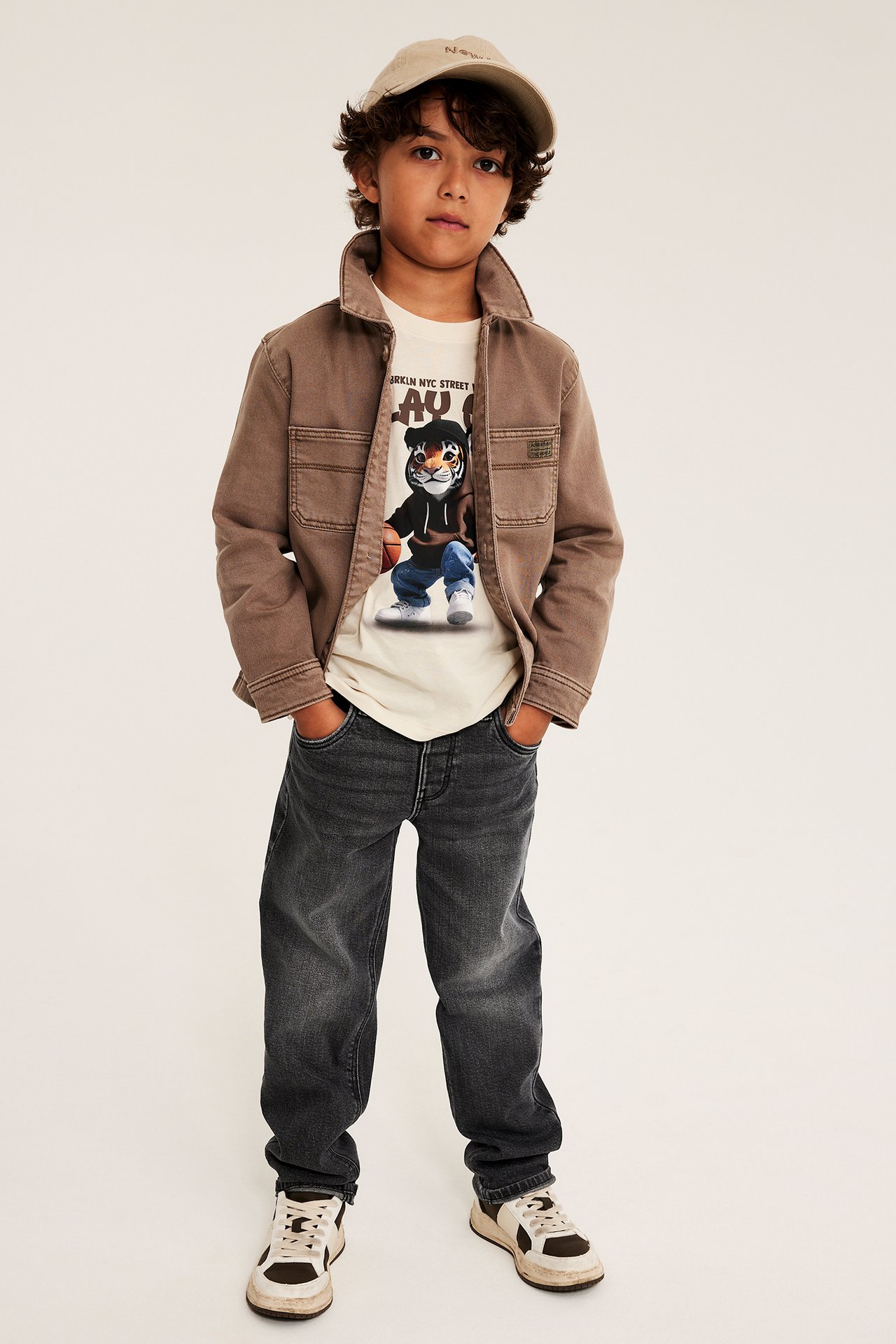 Pojke i brun skjortjacka, beige mönstrad t-shirt, mörkgrå jeans, sneakers och beige keps.