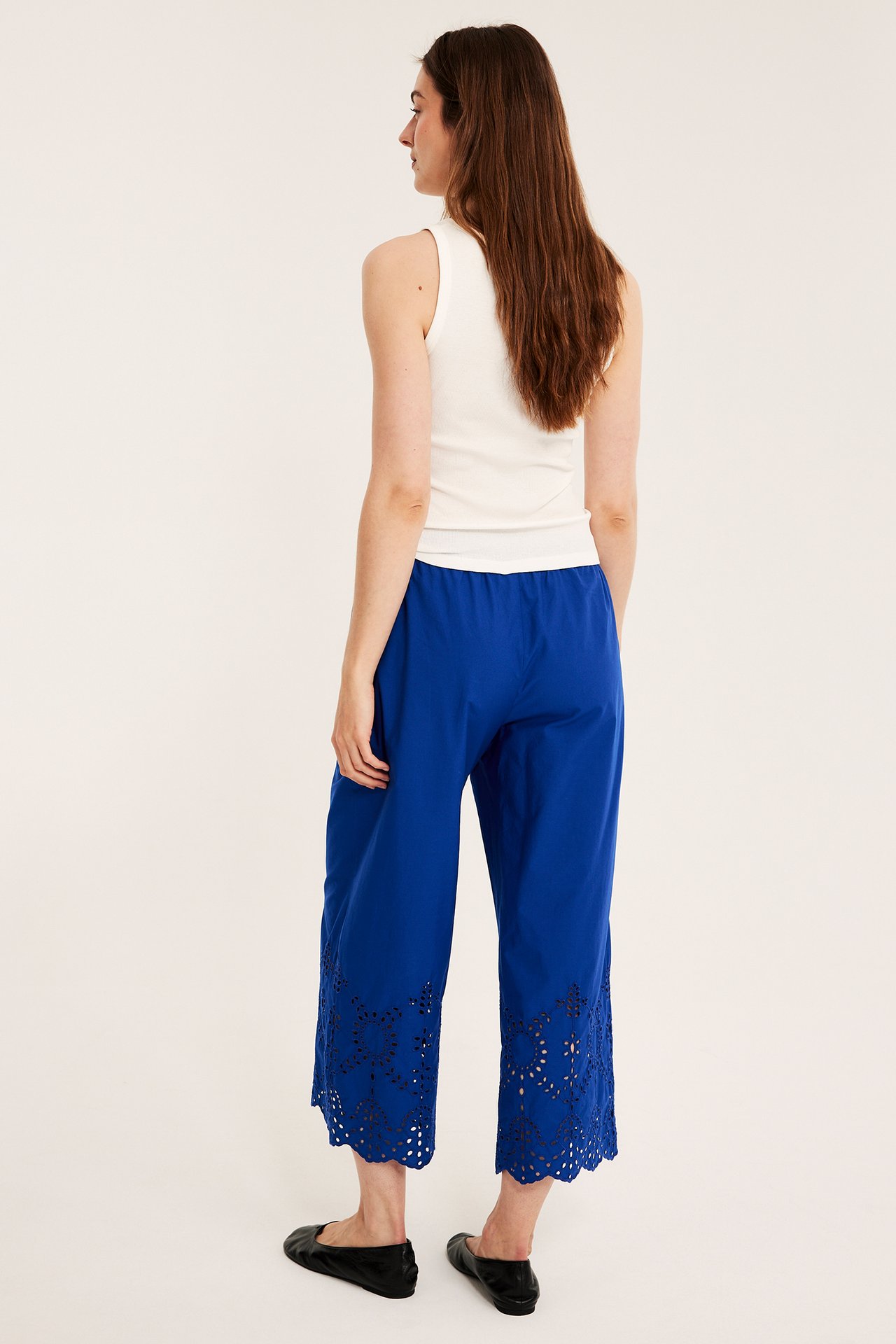 Naisen takakuva sinisissä leveissä puuvillaculottehousuissa, joissa broderie anglaise, valkoisessa topissa ja mustissa kengissä.