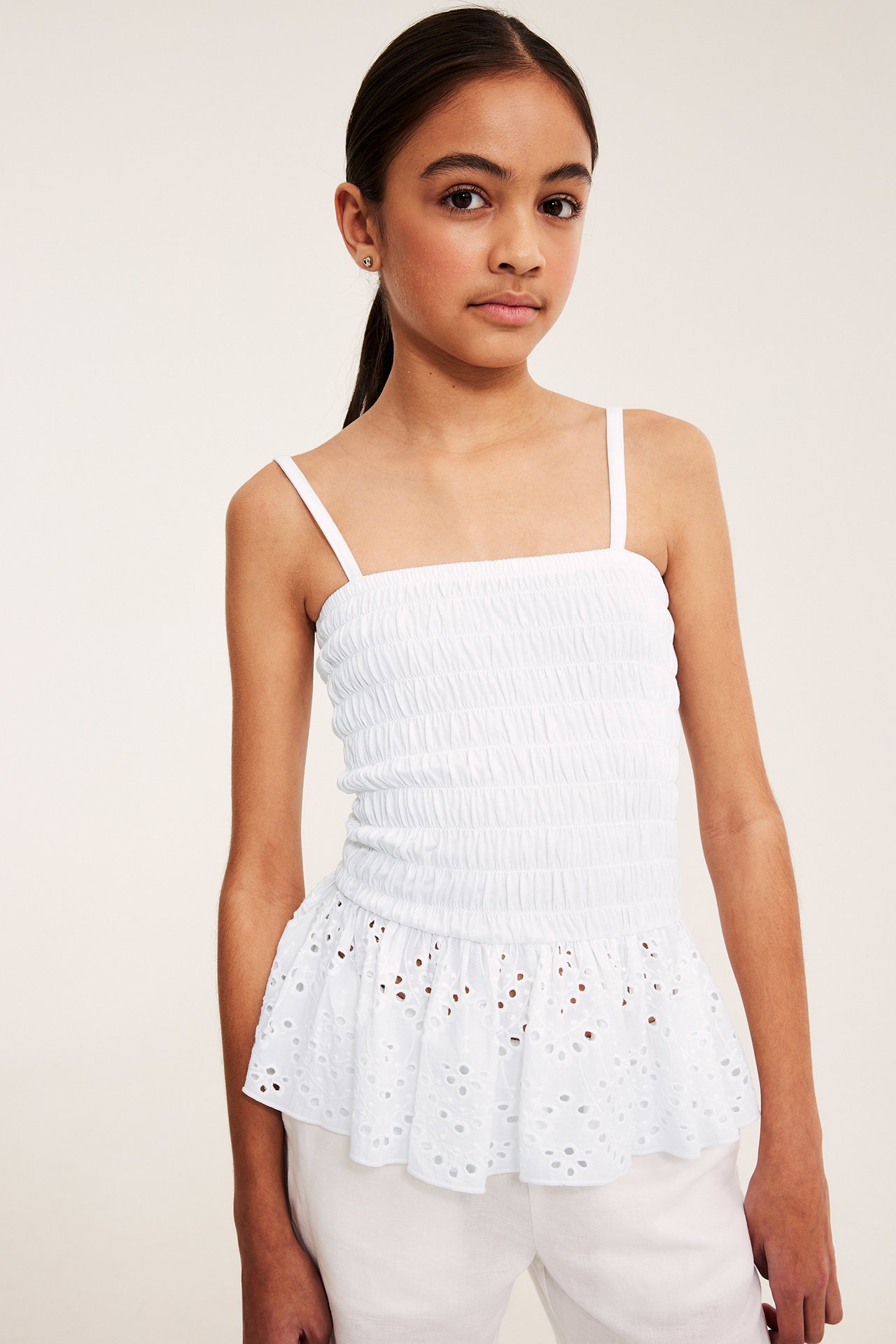 Framifrån: ett barn bär vit smockad topp med broderie anglaise-volang och vita byxor.