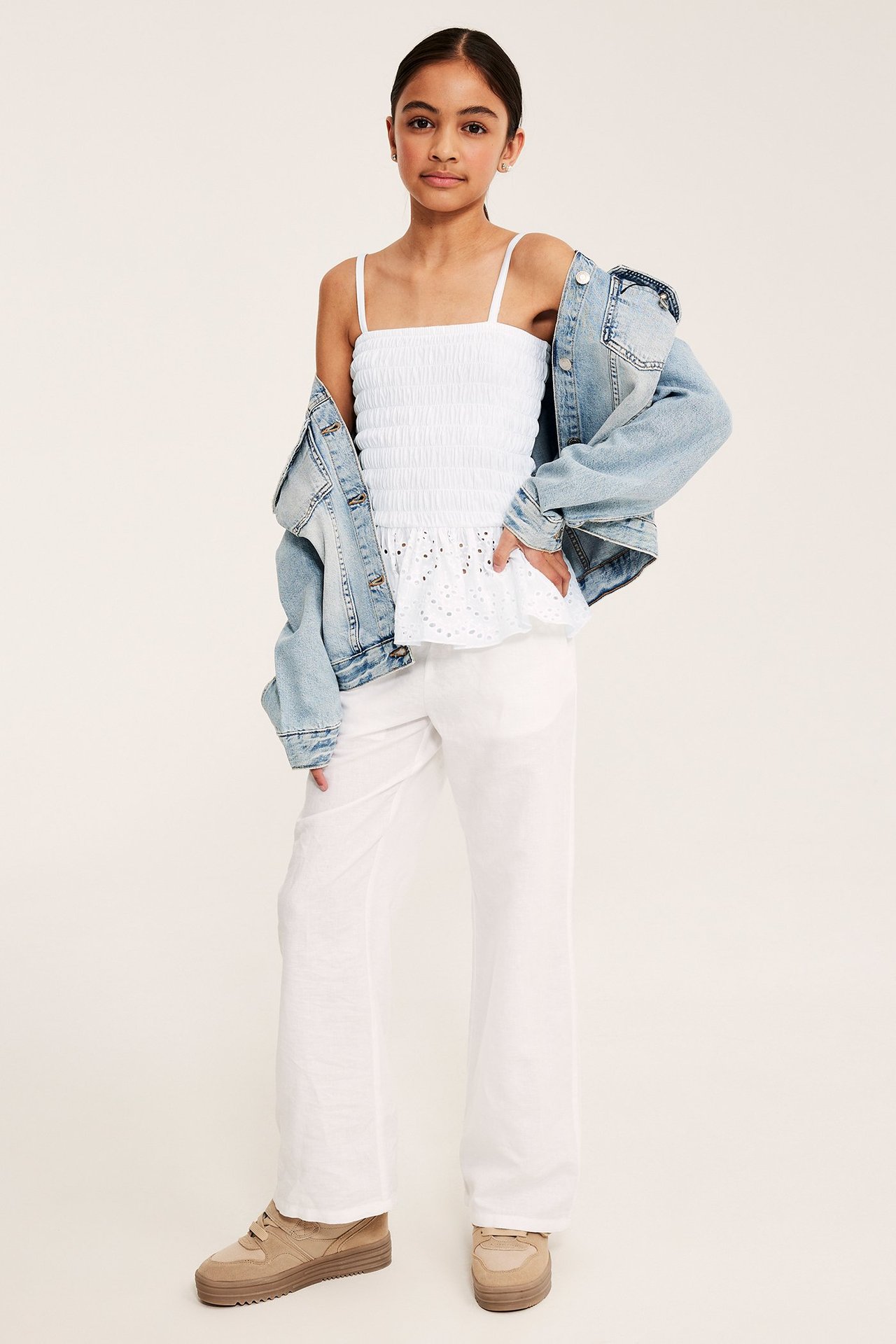 Frontvy av ett barn i vit smockad bandtopp med volang, ljusblå jeansjacka, vita vida byxor och beiga sneakers.