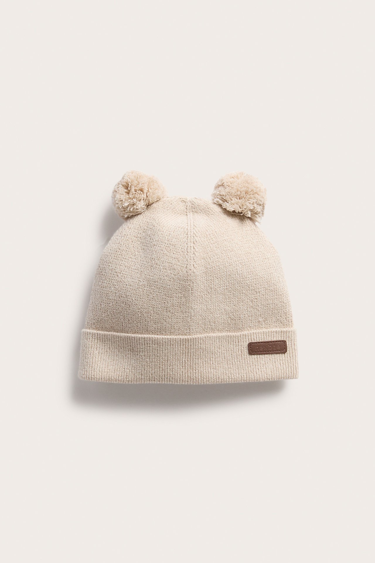 Frontbilde av en lys beige strikket baby lue med to dusker og en brun etikett.