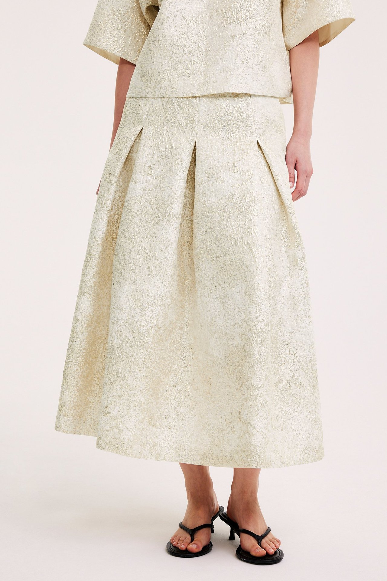 Frontbilde: kvinne i offwhite jacquardmønstret plissert skjørt, matchende topp og svarte flip-flop hæler.