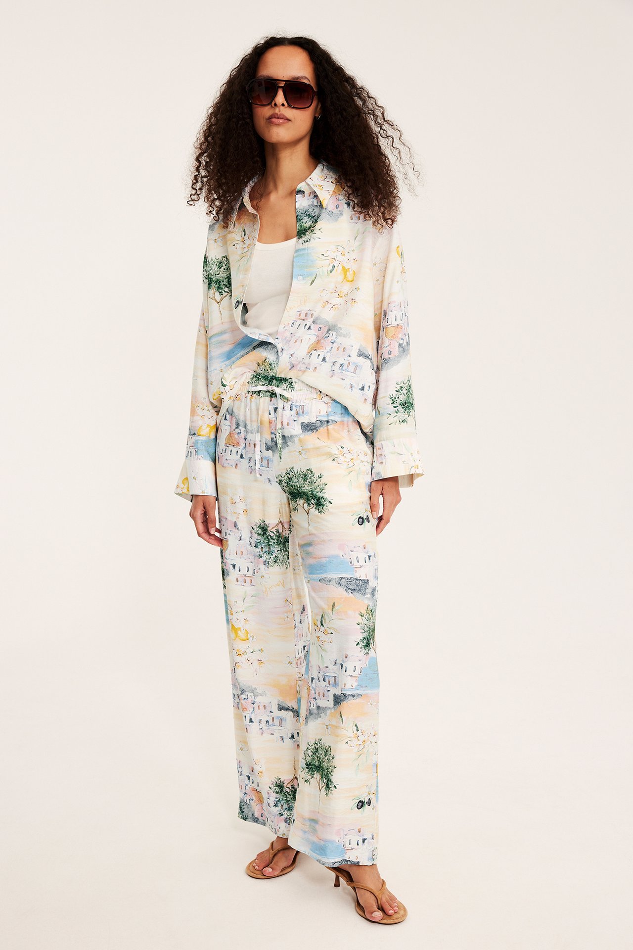 Frontvisning: Kvinne i offwhite mønstret vid viskosebukse med snøring, matchende skjorte, hvit topp, brune sandaler og solbriller.