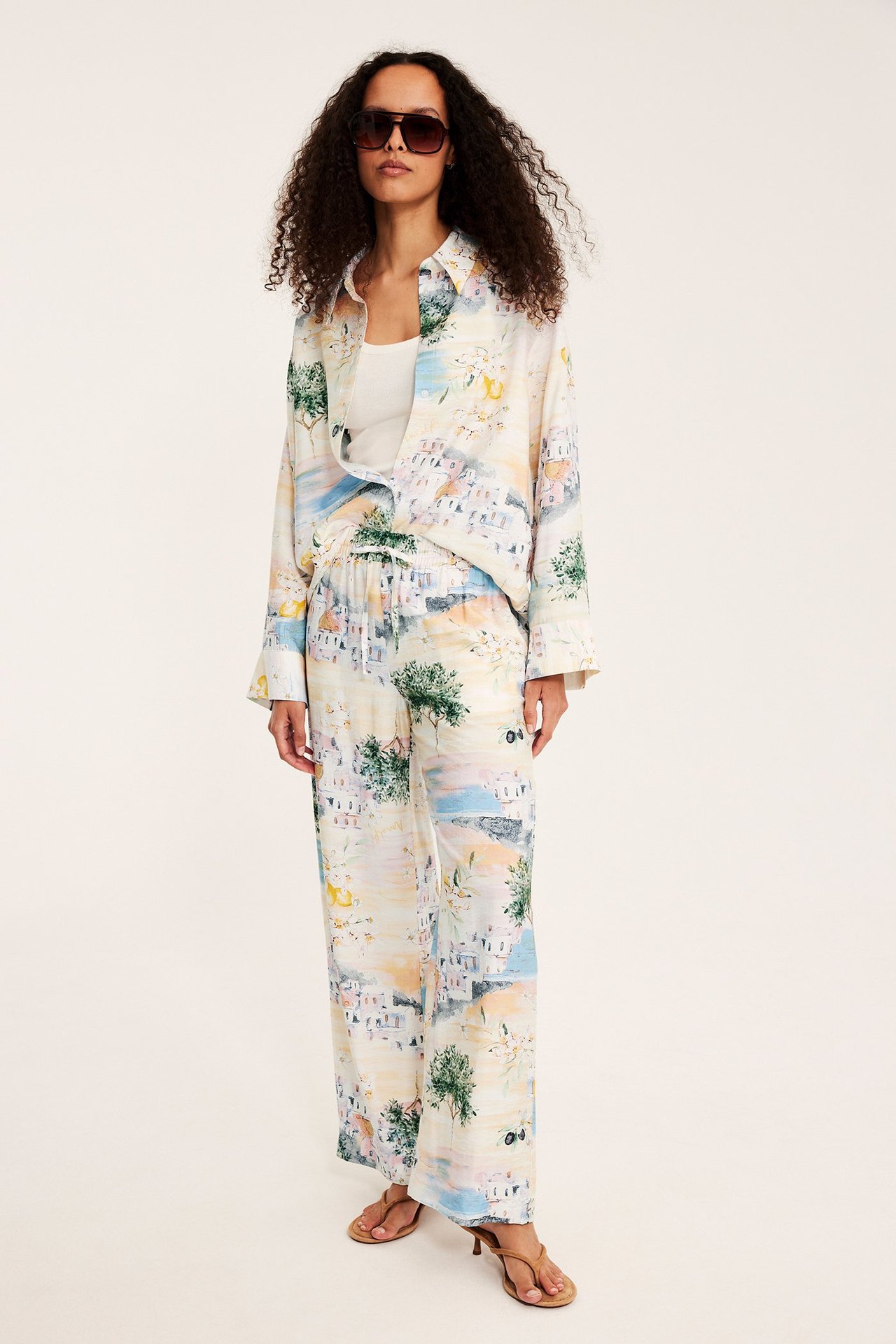 Framifrån: Kvinna i offwhite mönstrade vida byxor med dragsko, matchande skjorta, vitt linne, bruna sandaler och solglasögon.