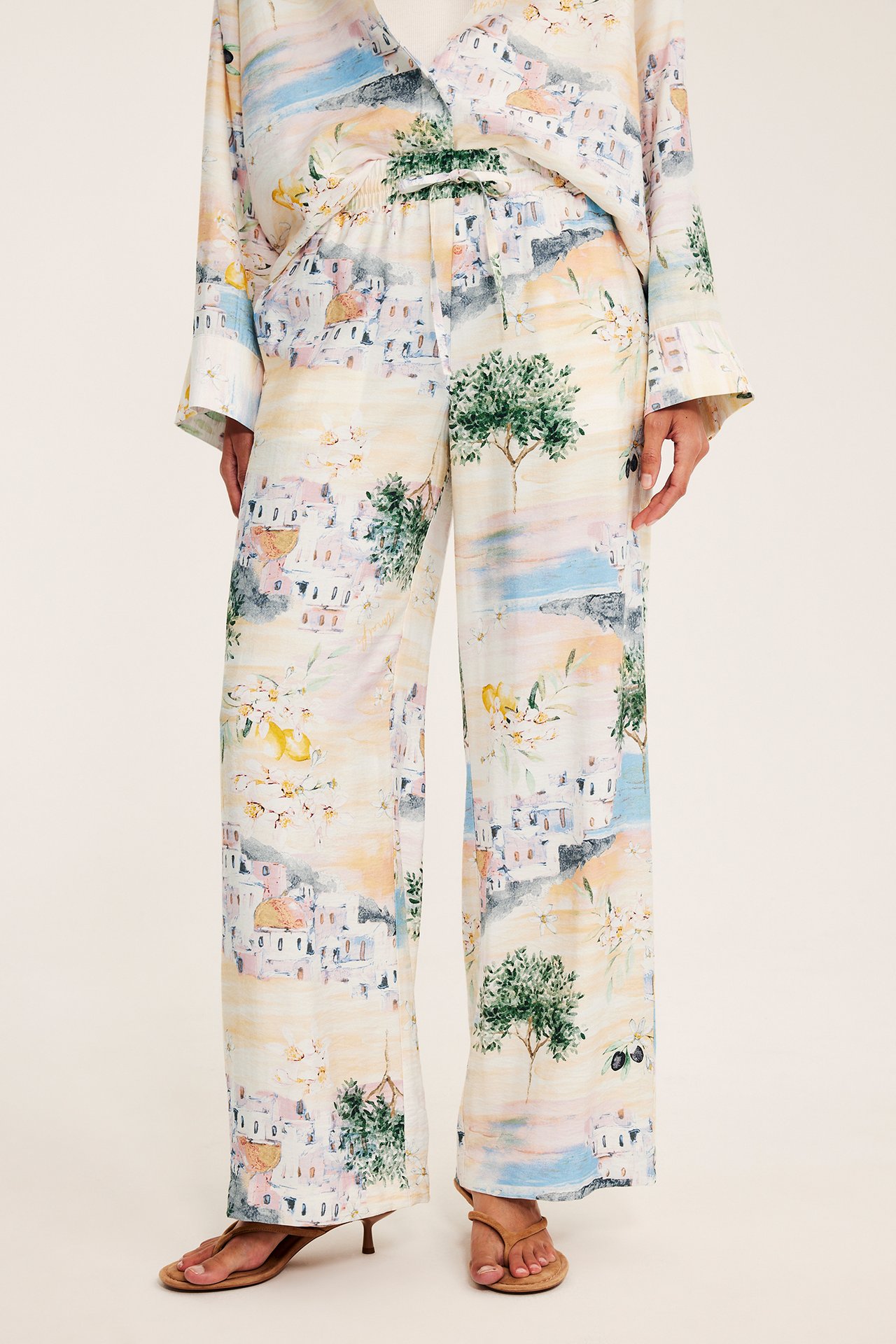 Framifrån: kvinna i vida offwhite viskosbyxor med Medelhavsmönster, matchande skjorta och bruna sandaler.