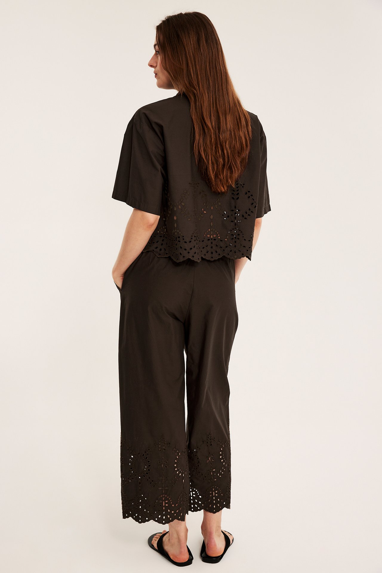 Bakfra kvinne i mørkebrune bomulls culottebukser med broderie anglaise, matchende topp og svarte sandaler.