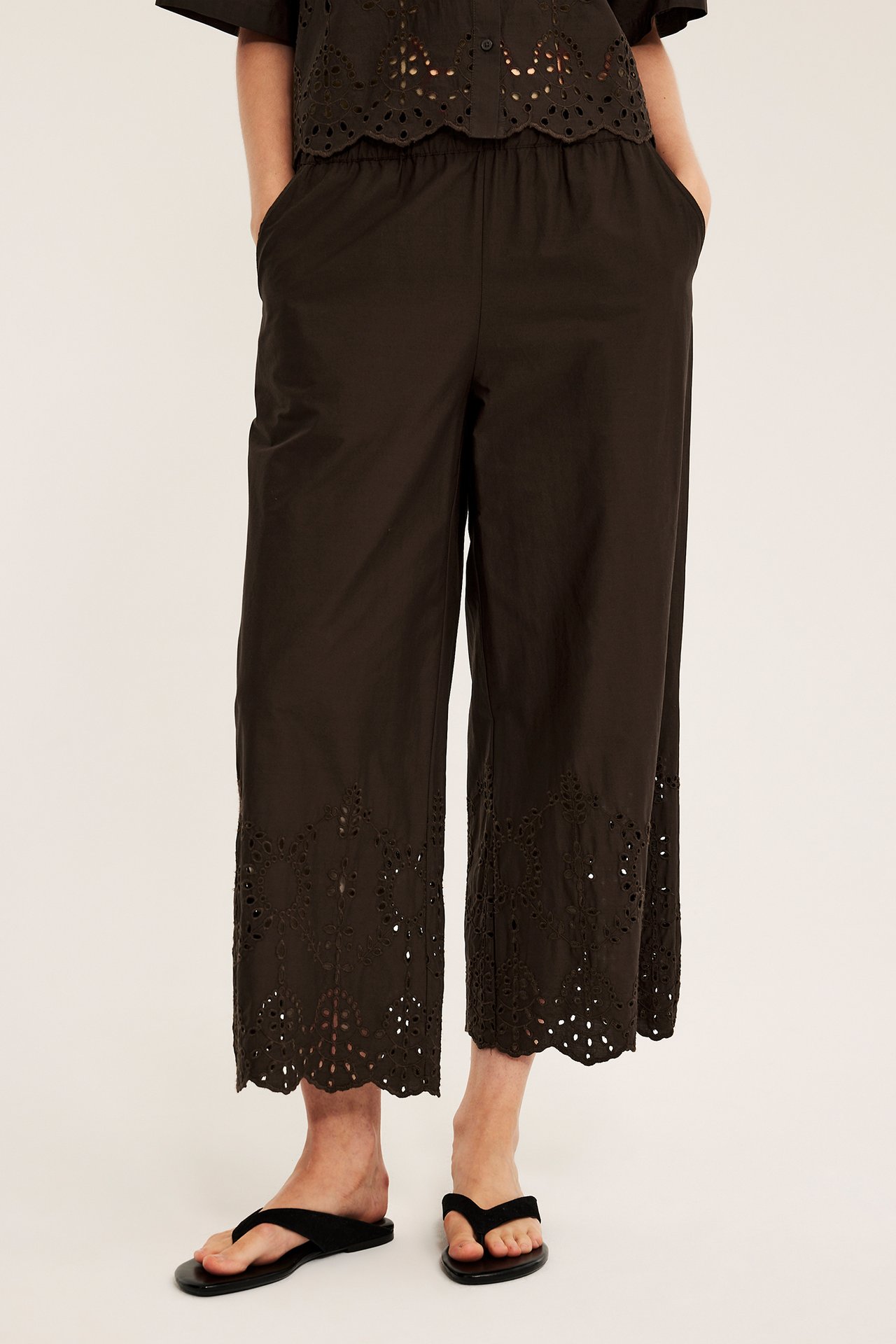 Widok z przodu: Kobieta w ciemnobrązowych bawełnianych spodniach z ażurowym haftem, pasującym topie i czarnych klapkach.