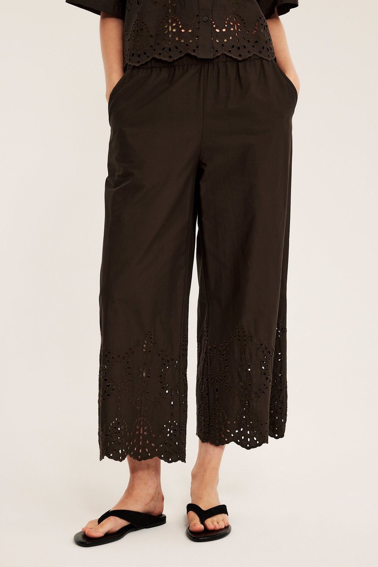 Edestä: Nainen tummanruskeissa puuvillahousuissa broderie anglaise -helmalla, sopiva toppi, mustat varvassandaalit.