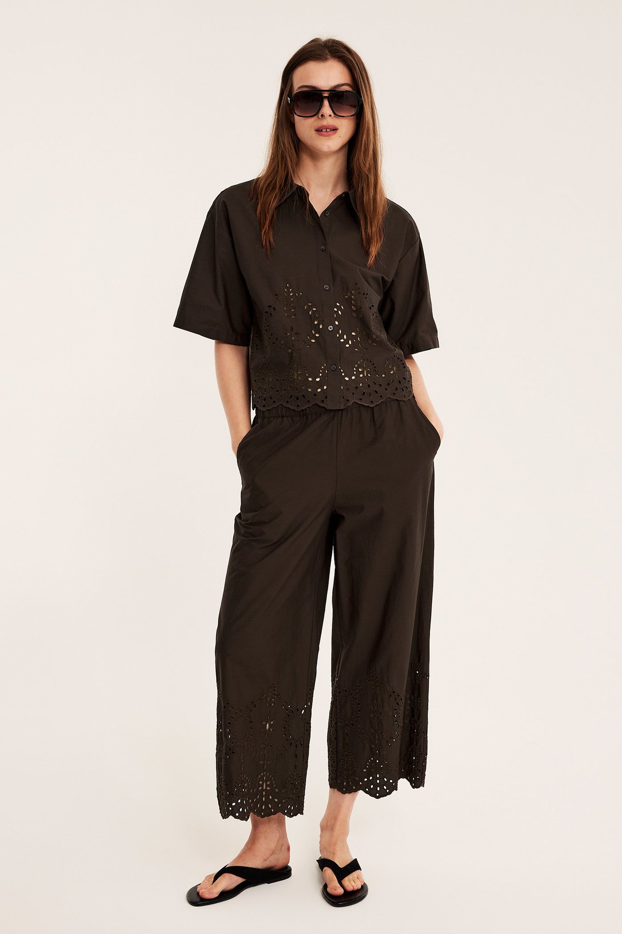 Edestä: nainen tummanruskeissa leveälahkeisissa puuvillahousuissa, joissa broderie anglaise, yhteensopiva lyhythihainen paita, mustat varvassandaalit ja aurinkolasit.